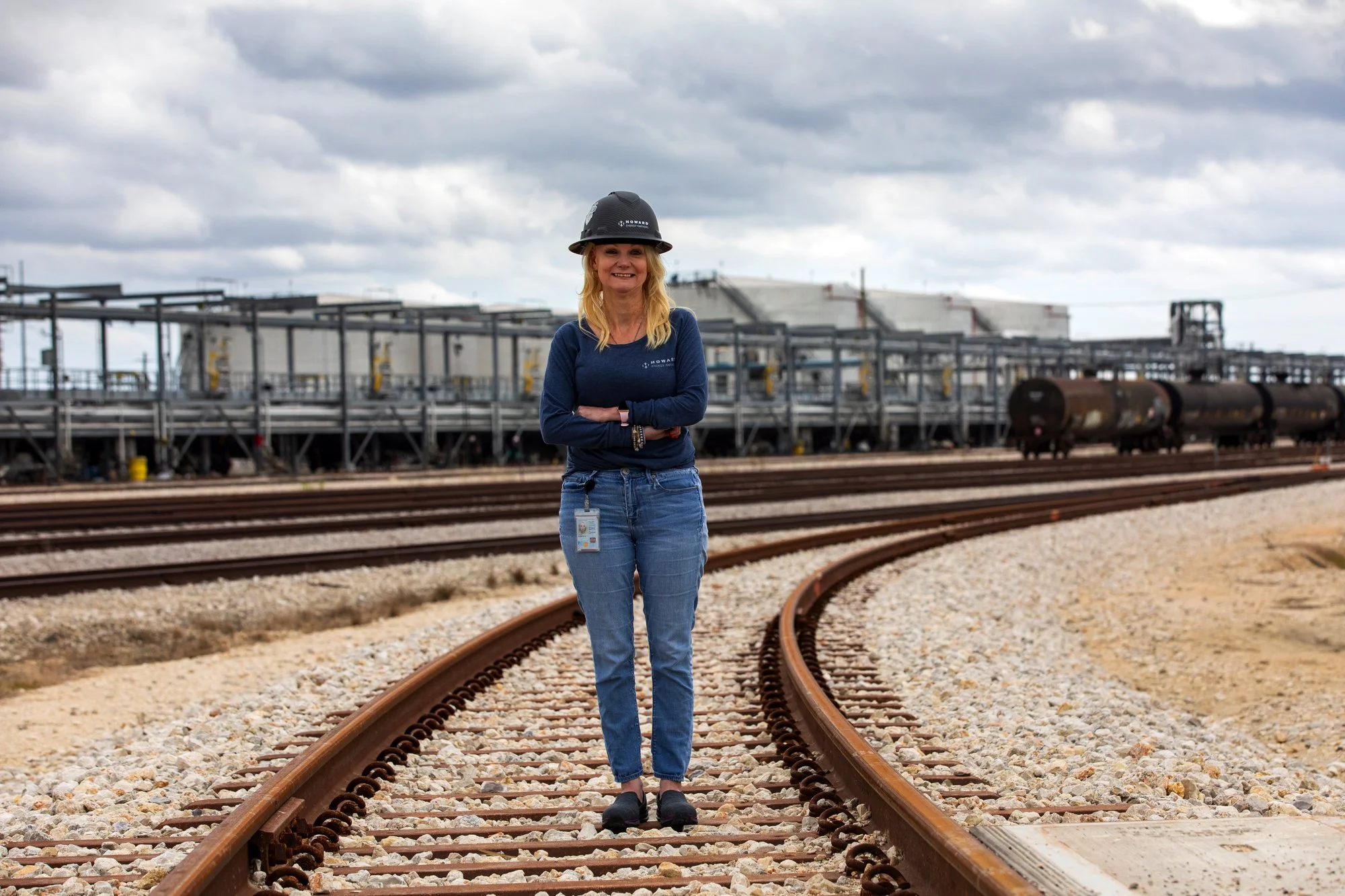 Woman standing on railroad tracks with industrial structures and train cars in the background on a cloudy day.