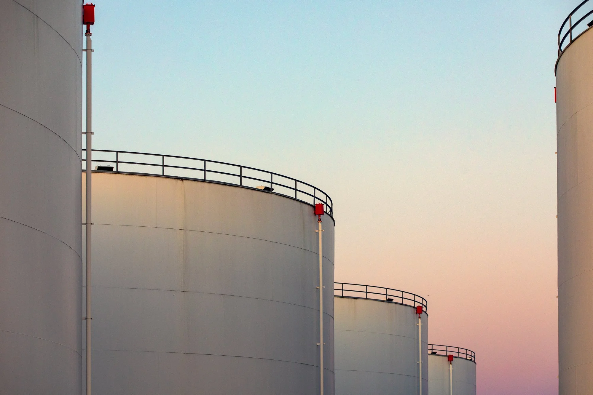 Large white industrial storage tanks with black railing on top, under a pale pastel sky.