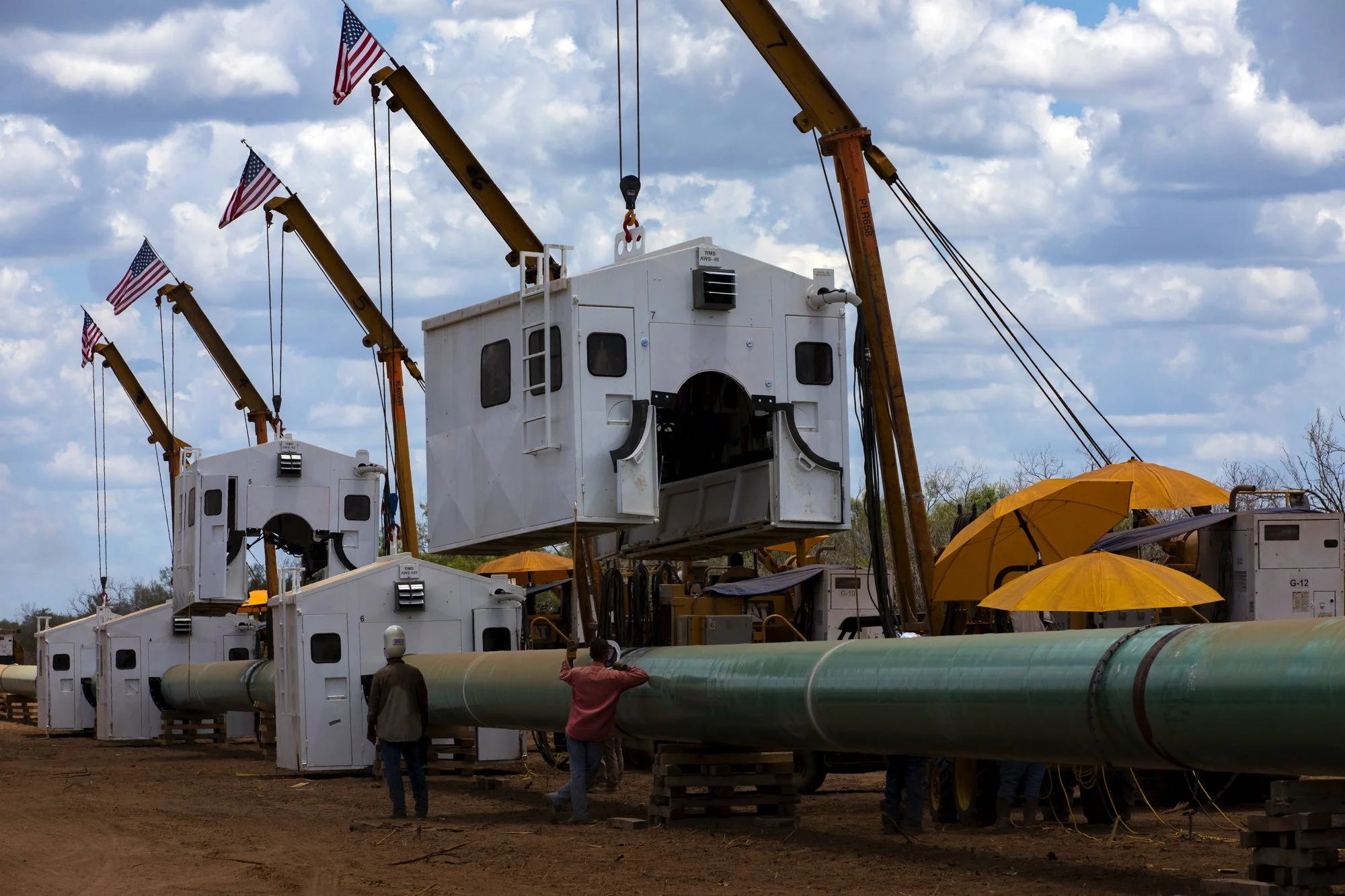 Construction site with large pipes, white control cabins, four yellow cranes with American flags, workers in safety gear, and yellow umbrellas.