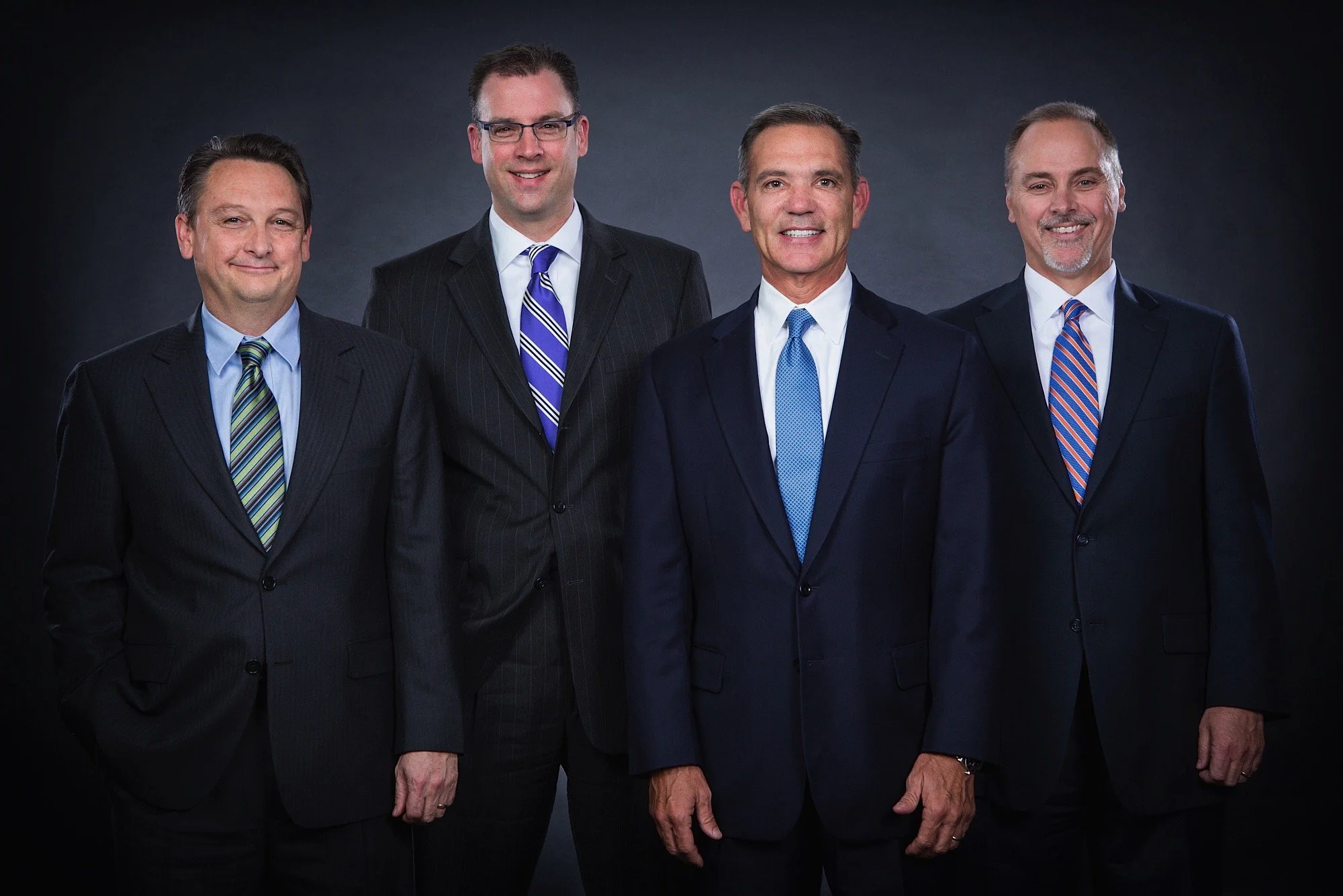 Five men in suits standing against a dark background, smiling at the camera.