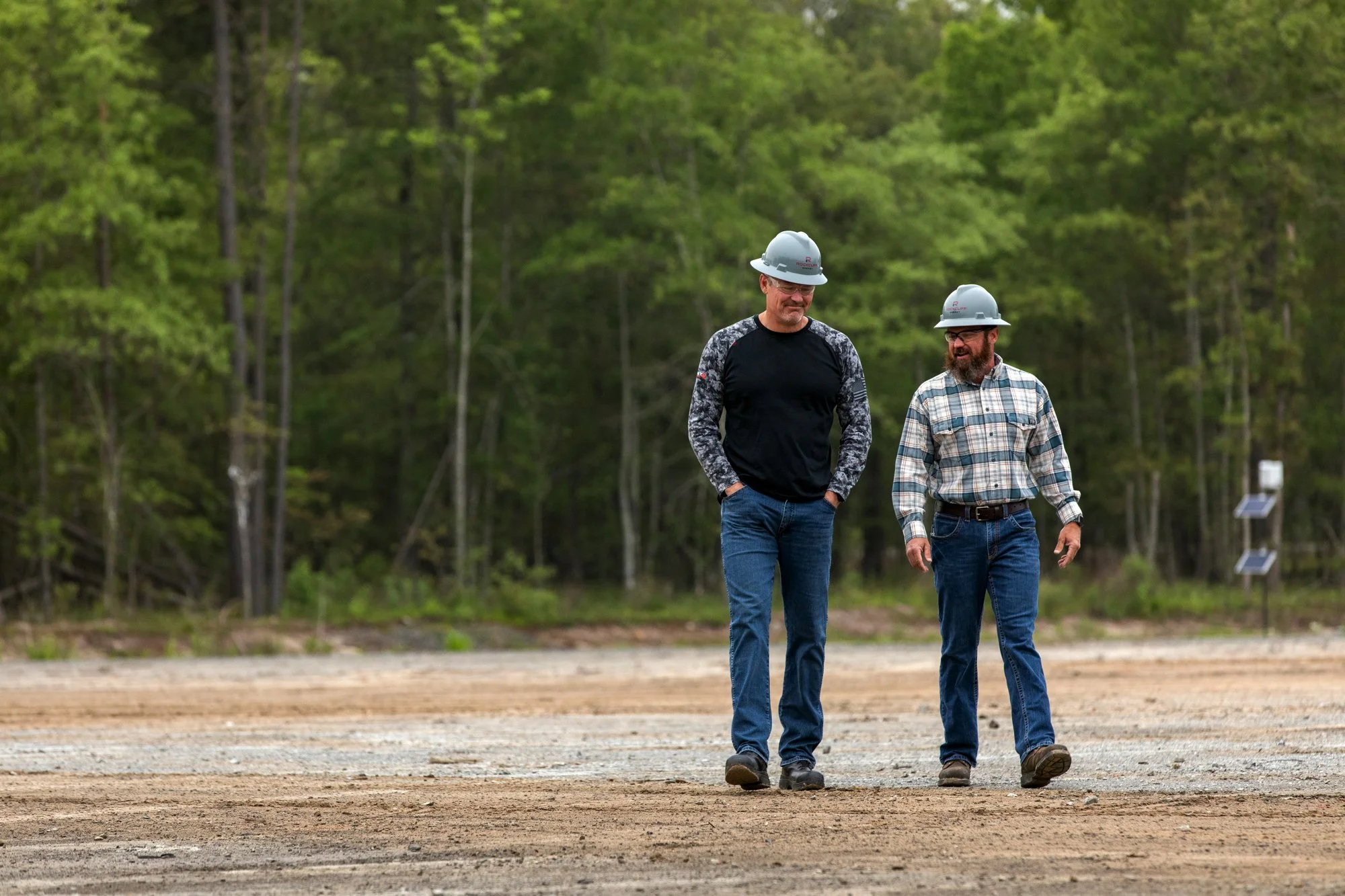 Two men wearing construction helmets walking and talking on a construction site with trees in the background.