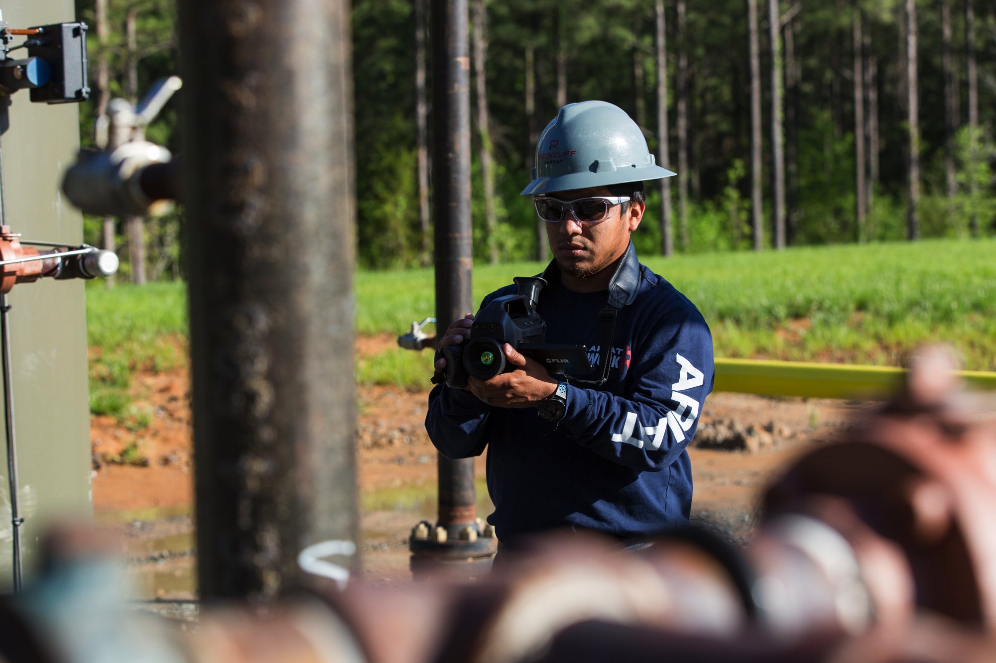 A male worker wearing a hard hat and safety glasses is inspecting or working on utility pipes outdoors in a green, wooded area.
