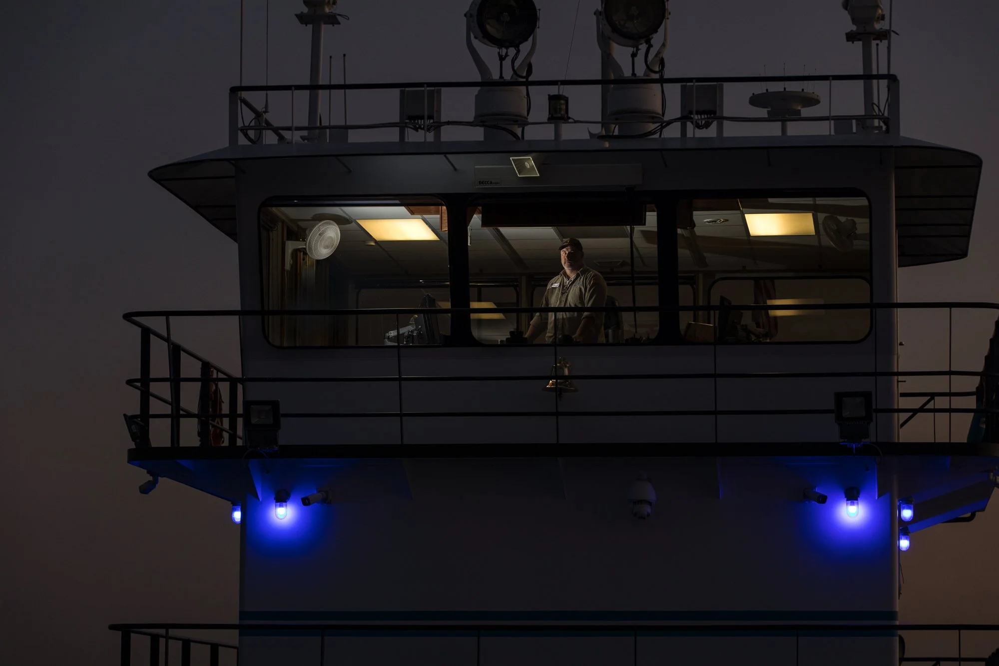A man standing inside the control room of a ship's bridge, illuminated by interior lighting, with windows showing the dark sky outside, and blue exterior lights below.