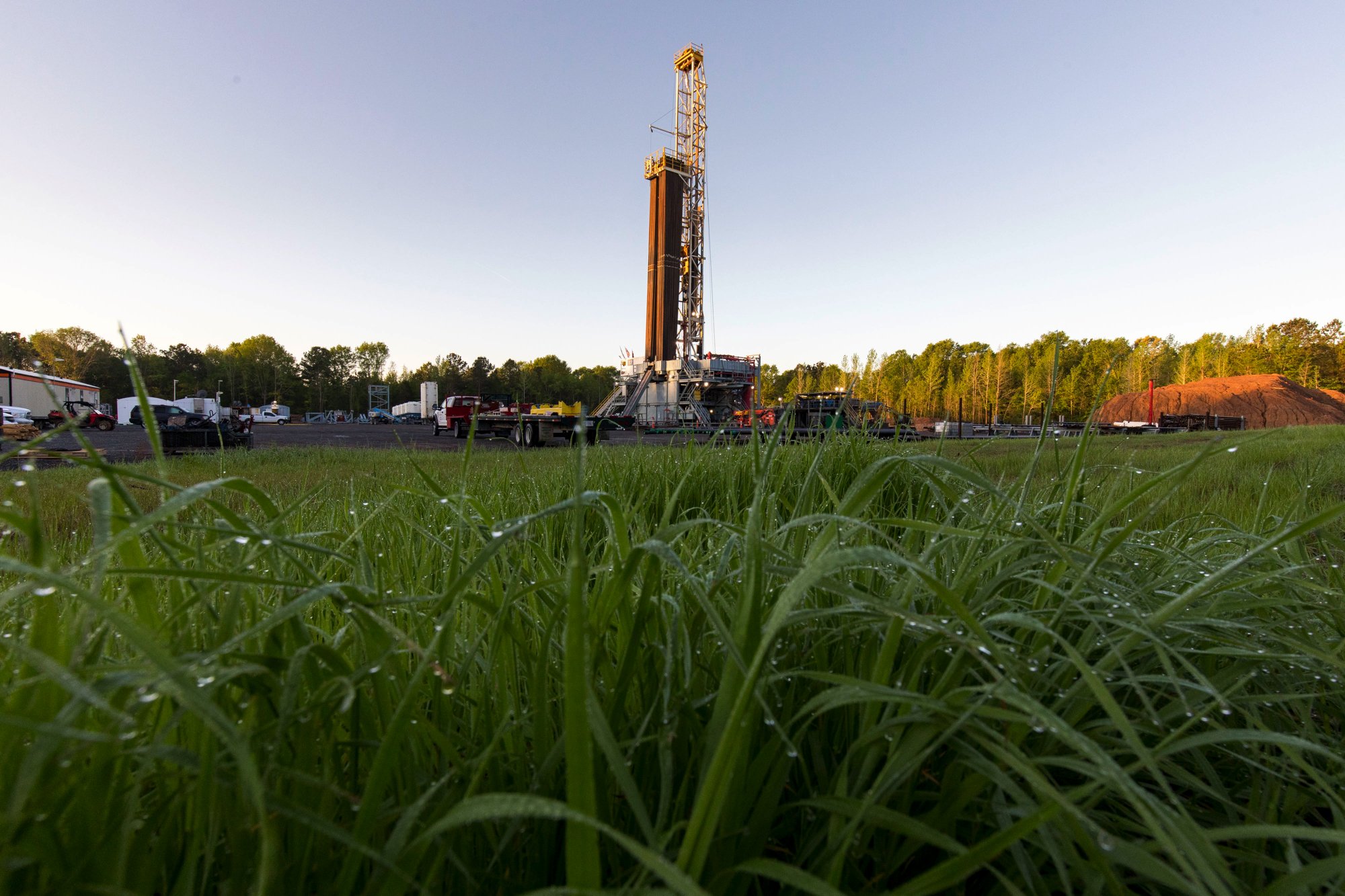 Drilling rig in a grassy field with various parked vehicles and equipment in the background under a clear sky.