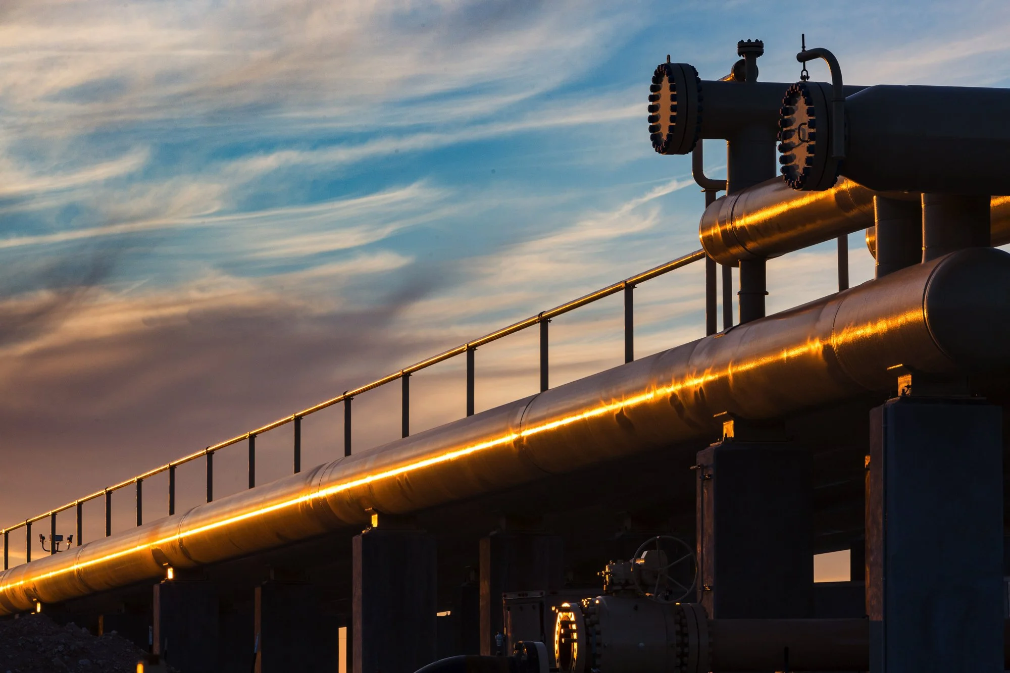 Oil or gas pipeline with large pipes and valves, reflecting sunlight at sunset or sunrise.