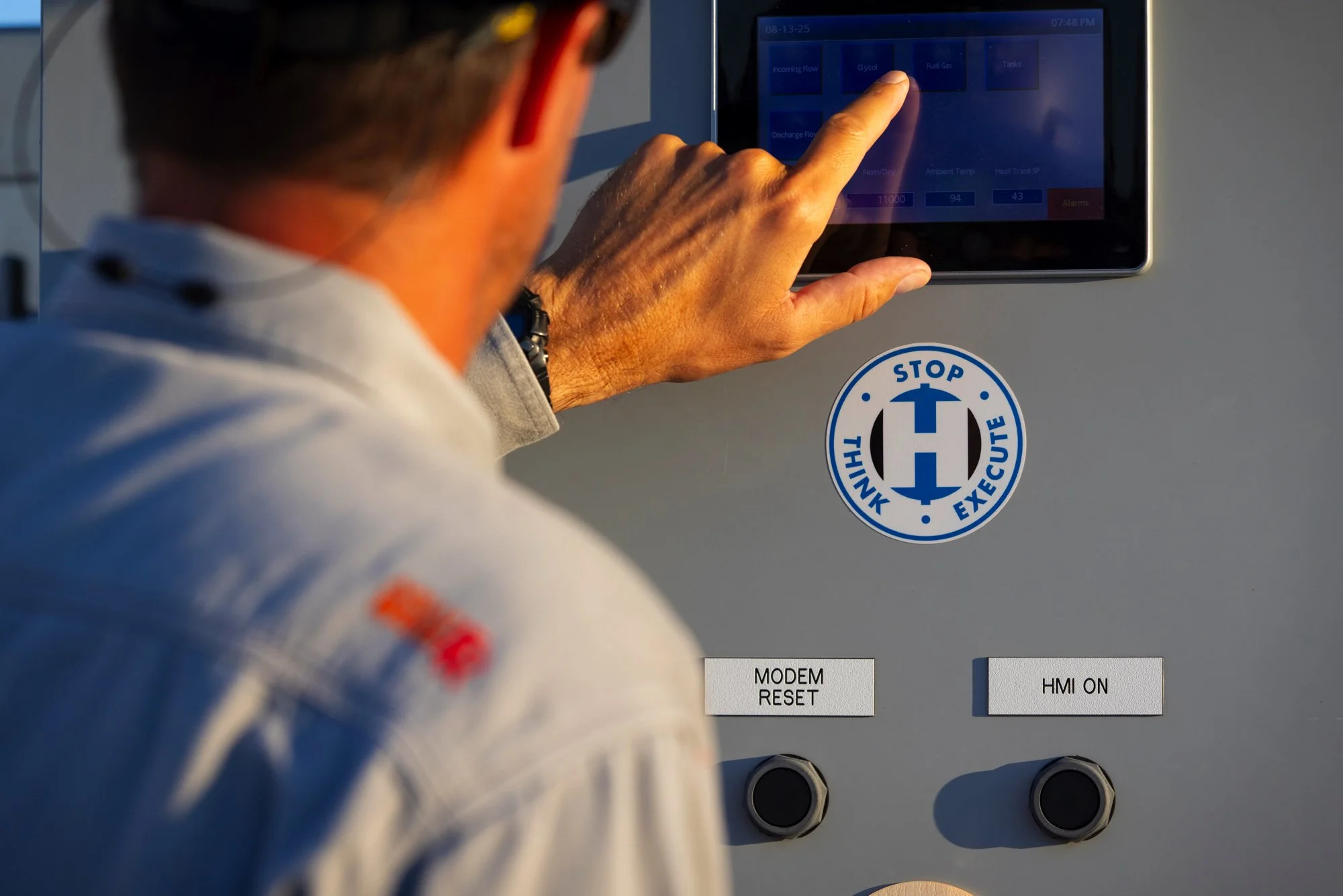 A person in a white racing suit touching a touchscreen control panel with labels such as 'Gycol,' 'Fuel Gas,' and 'Tanks.' There is a circular sticker with the words 'Stop Think Execute' and a graphic resembling a steering wheel or a directional symb