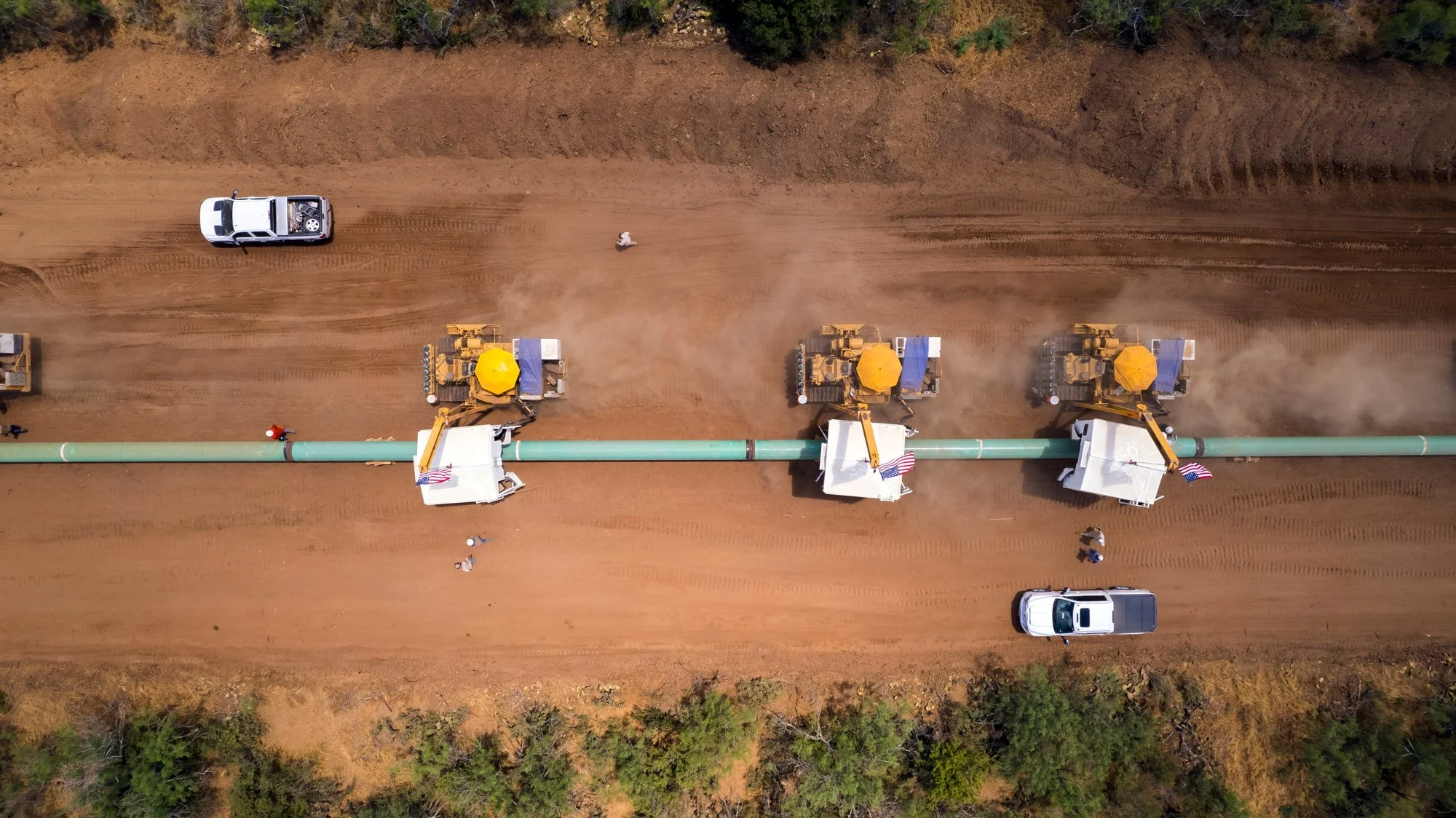 An aerial view of a pipeline construction site with three large machinery vehicles working along a large pipeline, flanked by trees and dirt roads, with a few workers visible.
