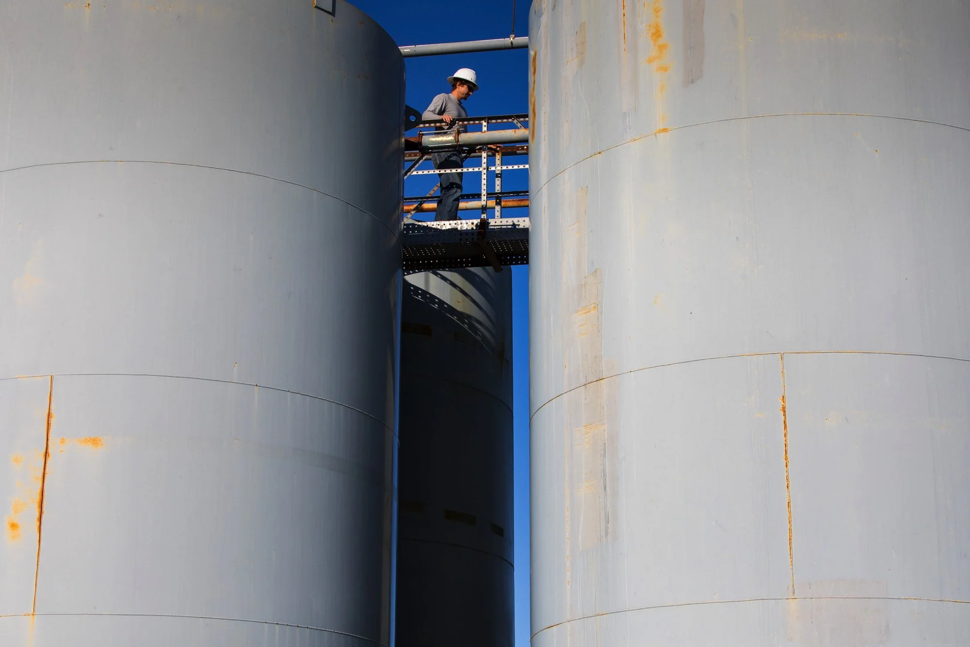 A worker in a hard hat standing on a metal walkway between two large gray industrial storage tanks or silos.