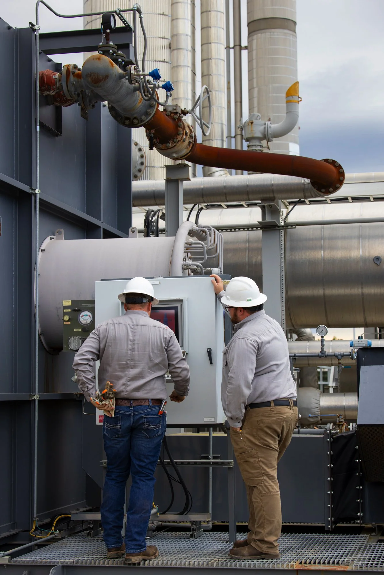 Two workers wearing hard hats and work clothes inspecting a control panel on industrial equipment at an outdoor facility with pipes and large storage tanks.