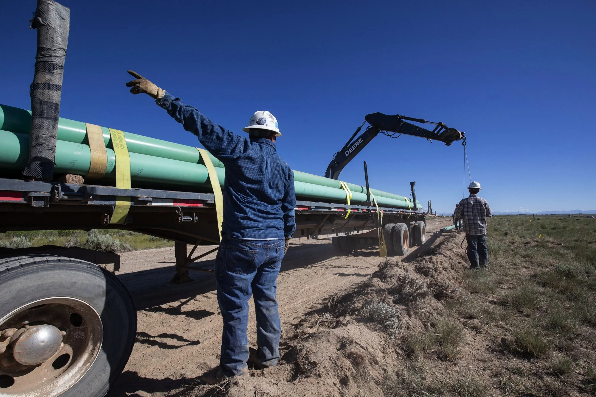 Two construction workers wearing safety helmets work on a pipeline installation in a rural area. One worker directs the operation, while the other stands near the pipeline with a excavator in the background.
