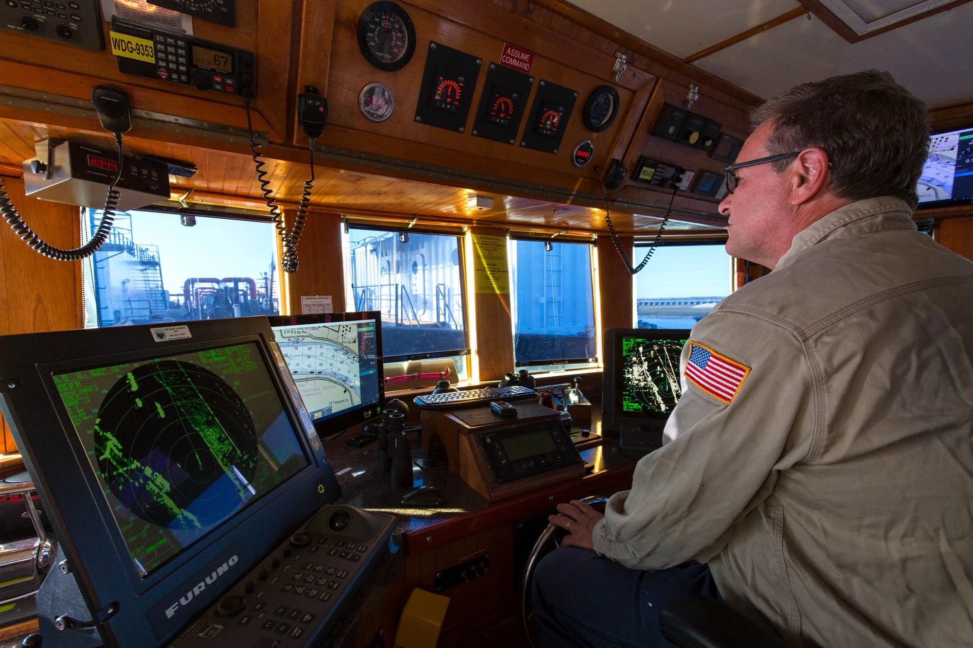 A ship captain operating navigation equipment in the ship's control room, with radar, communication devices, and monitors displaying navigational charts and radar images.