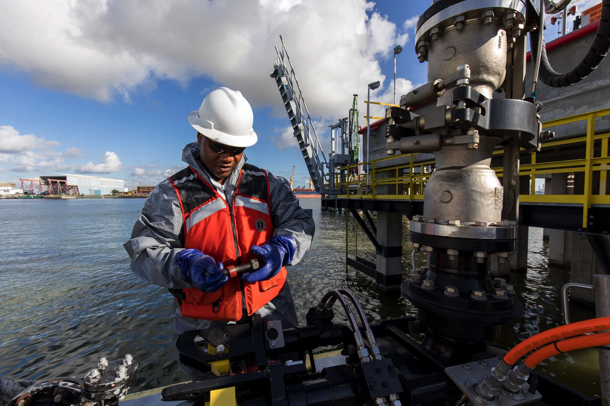 A worker wearing a white safety helmet, sunglasses, an orange safety vest, and blue gloves inspecting equipment near the water's edge on an industrial platform, with a large industrial structure, water, and a partly cloudy sky in the background.
