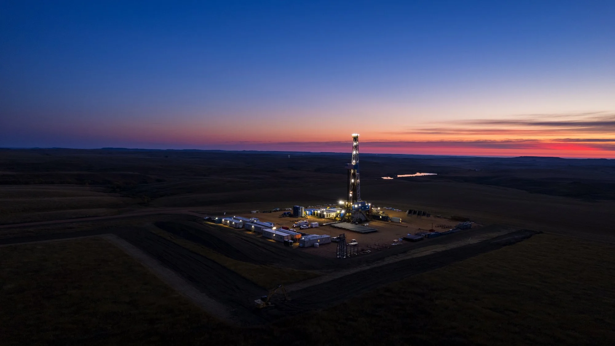 An oil drilling rig illuminated at twilight in a flat, open landscape with a body of water in the distance.