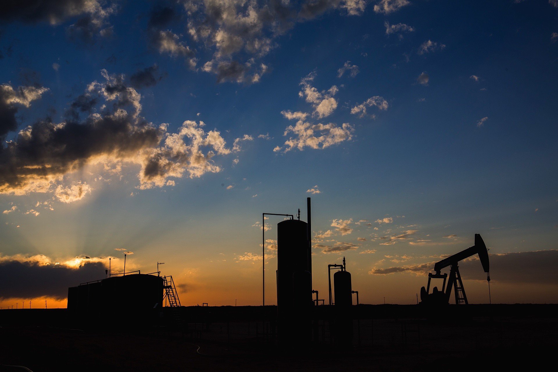 Silhouettes of oil drilling equipment and storage tanks against a sunset sky with clouds.