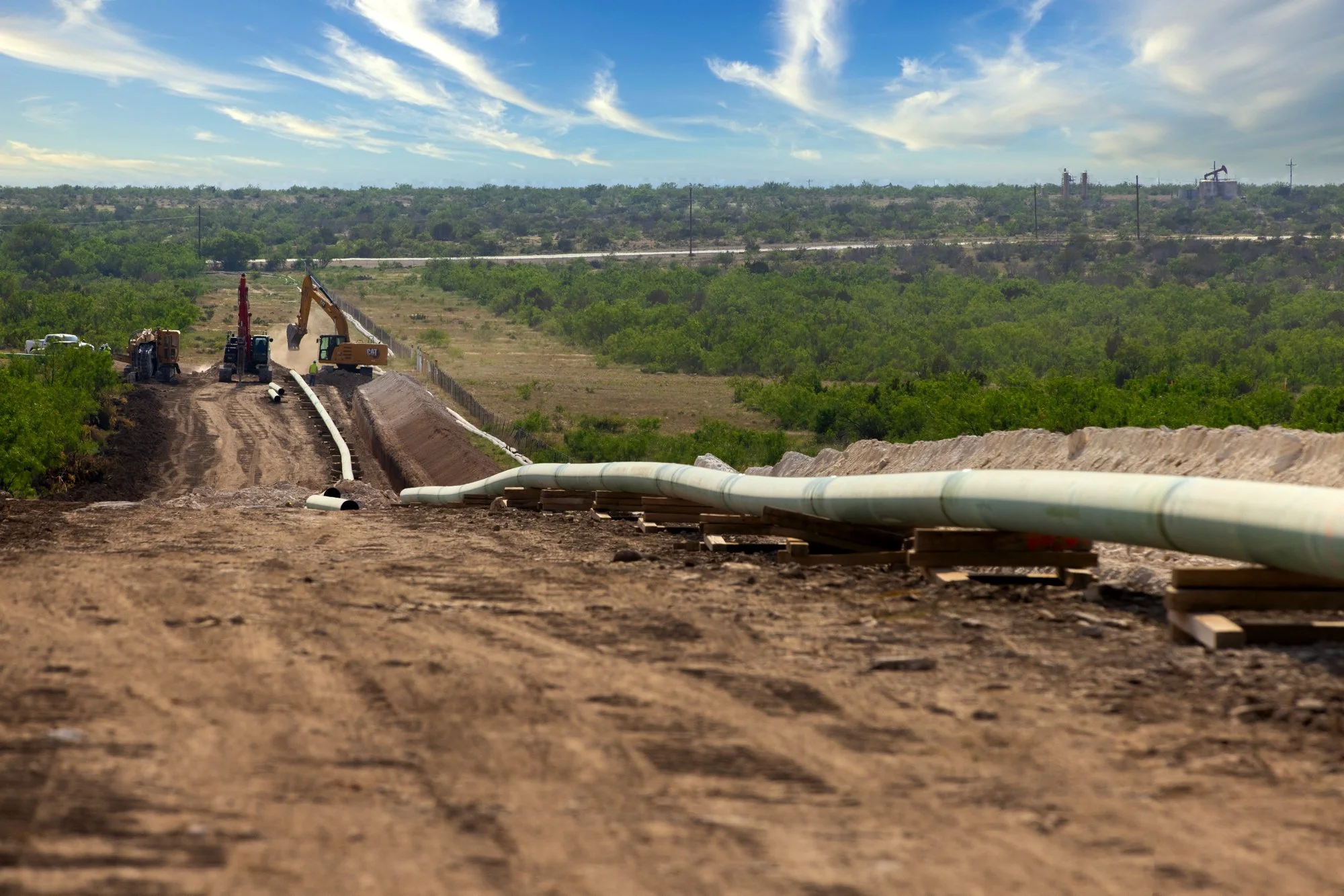 Construction workers and machinery installing underground pipes in a rural area with green trees and blue sky.