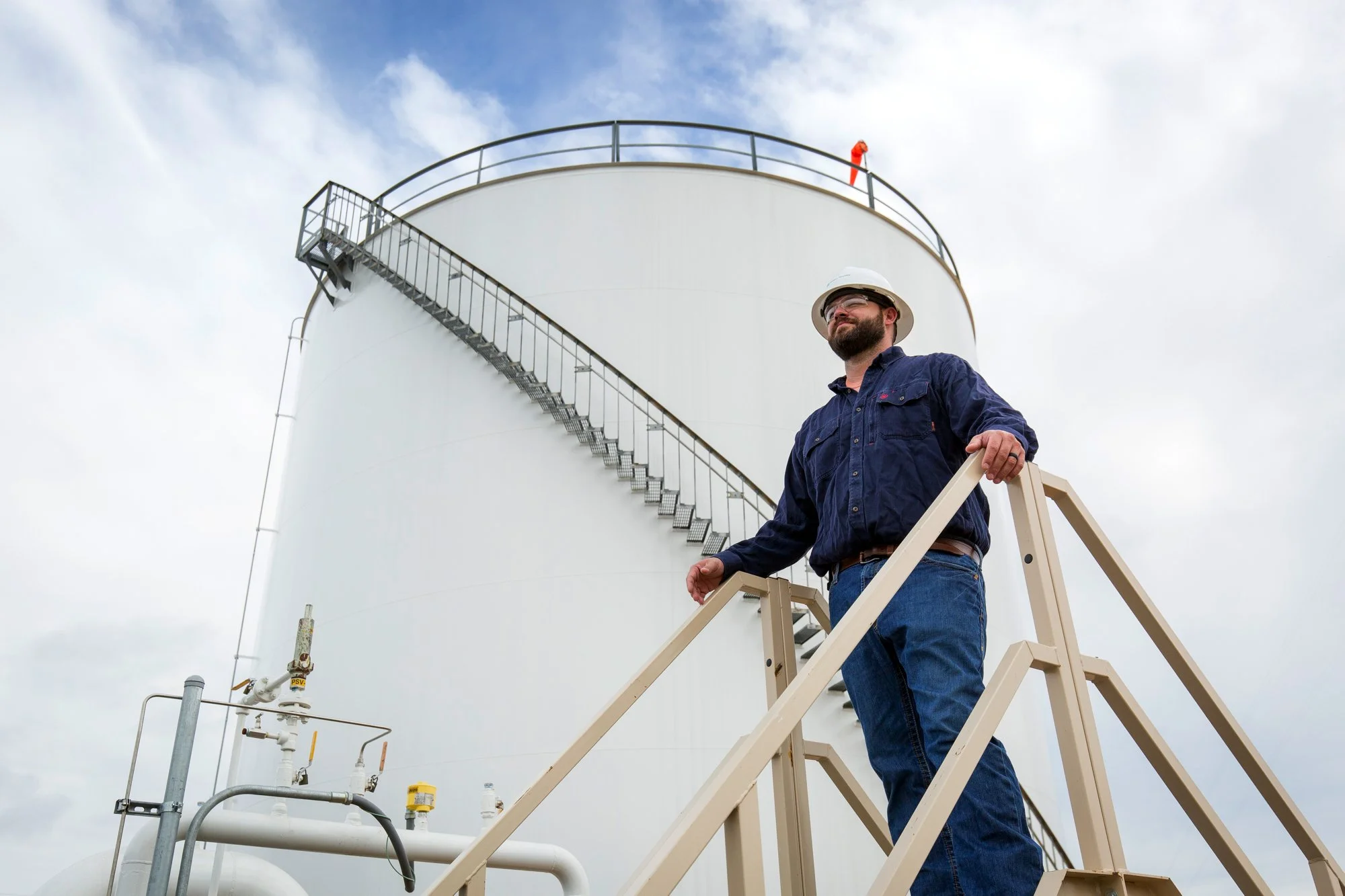 A man wearing a hard hat and safety glasses stands on a staircase at an industrial tank facility, with pipes and valves nearby under a cloudy sky.