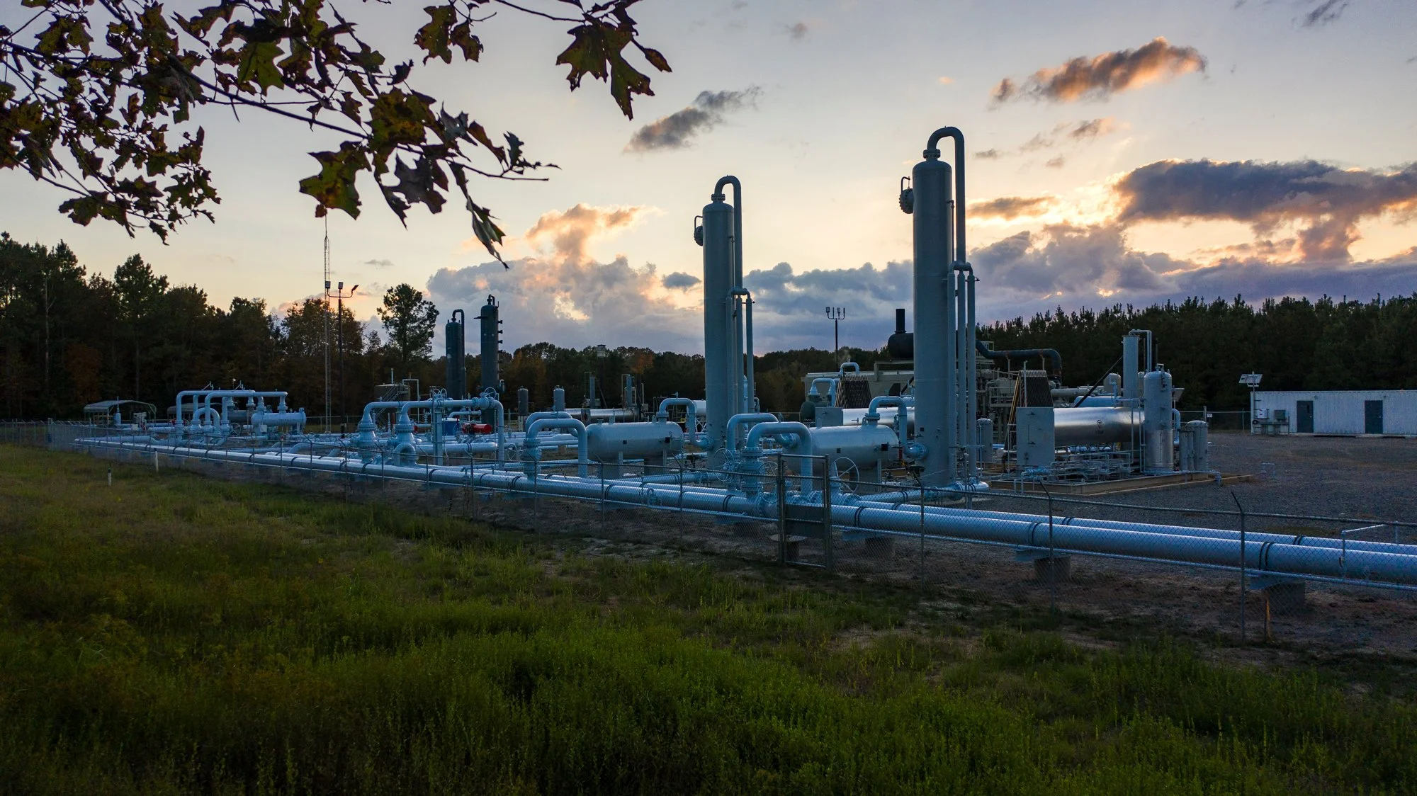 Industrial gas processing facility with pipelines and equipment at sunset, surrounded by trees and a grassy area.