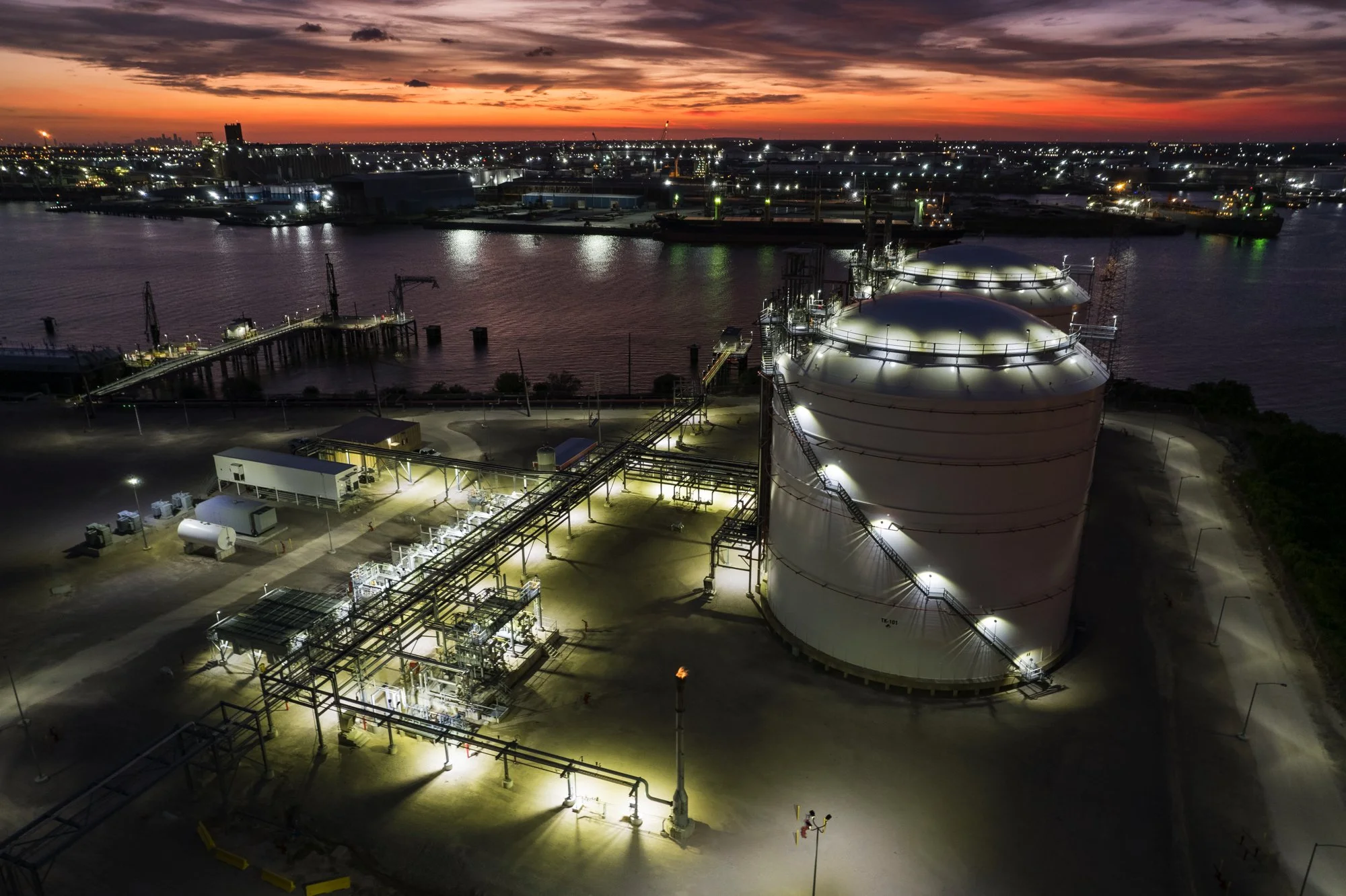 Nighttime view of an industrial port area with large white storage tanks illuminated by lights, pipelines, and equipment in the foreground, with a body of water and city skyline with buildings and lights in the background, under a sunset sky with pur