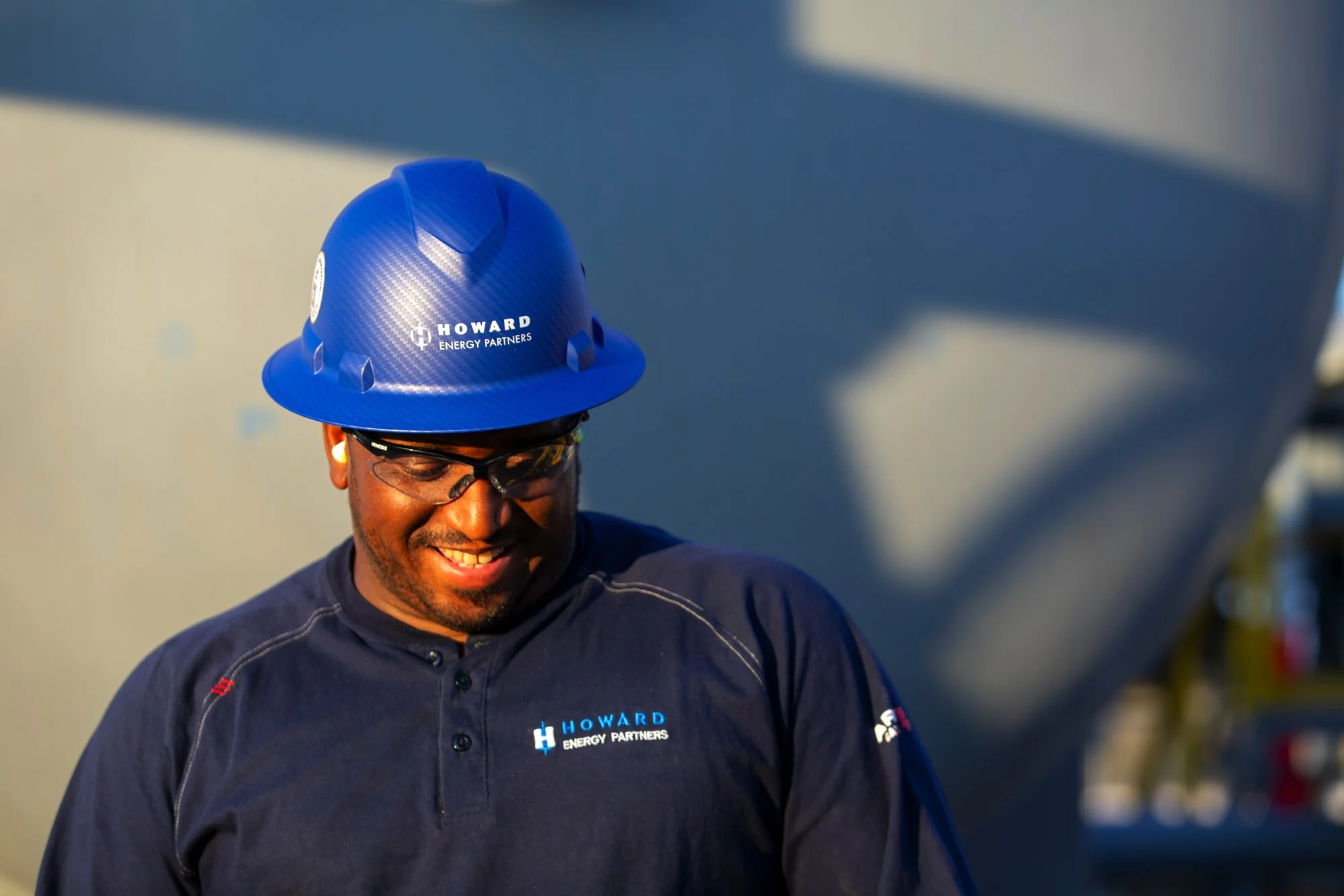 A man wearing safety glasses and a navy blue hard hat with 'Howard Energy Partners' logo, smiling and looking down, outdoors in sunlight.