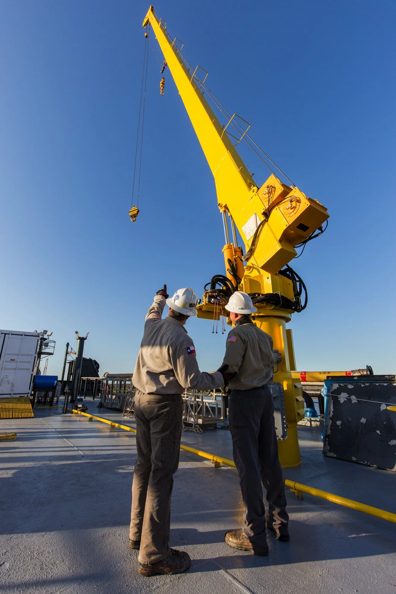 Two workers in safety gear shake hands on a deck with a large yellow crane overhead.