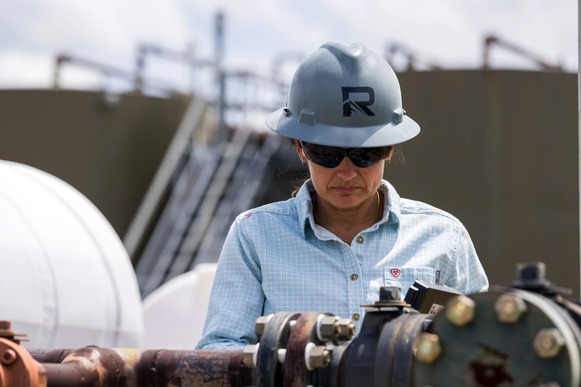 A woman wearing a hard hat, sunglasses, and a light blue button-up shirt inspecting large industrial pipes outdoors.