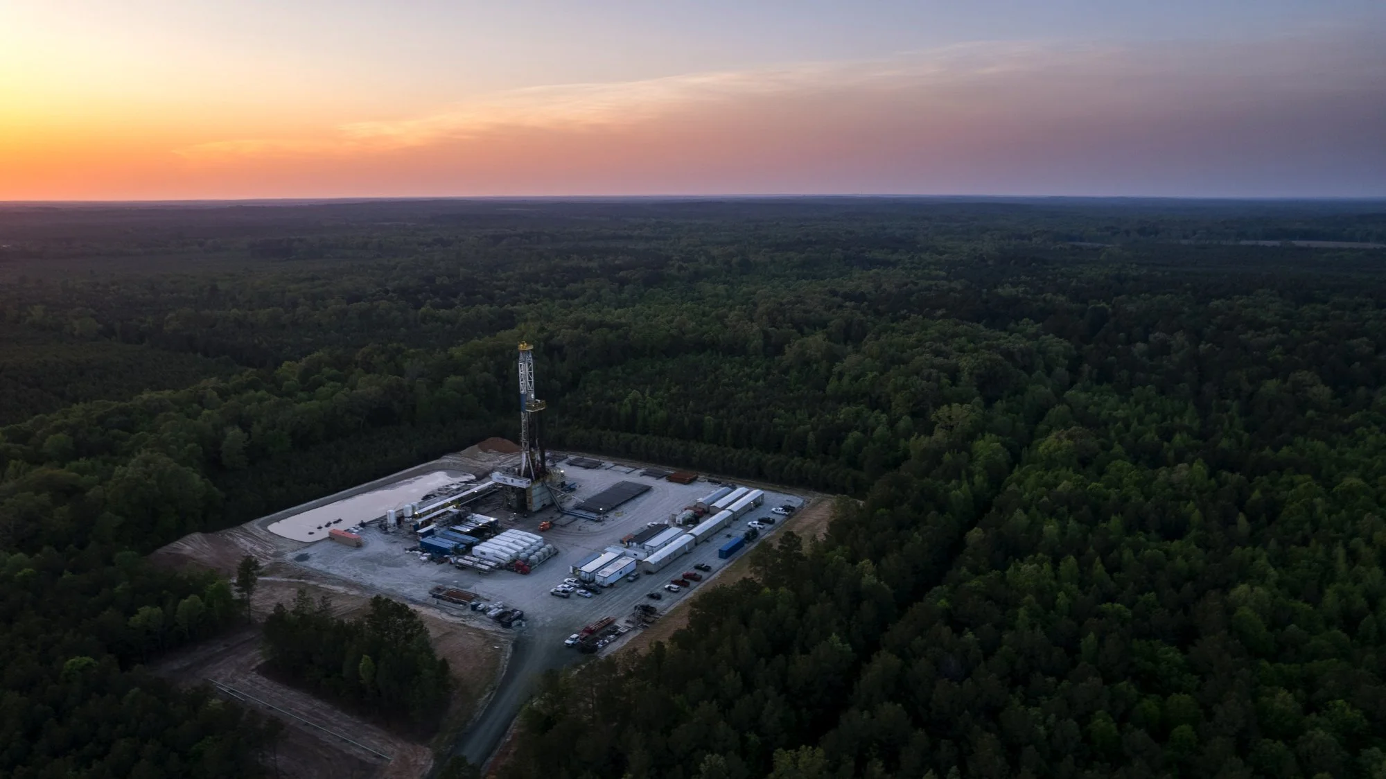 An oil drilling rig with surrounding equipment and vehicles in a forested area at sunset.