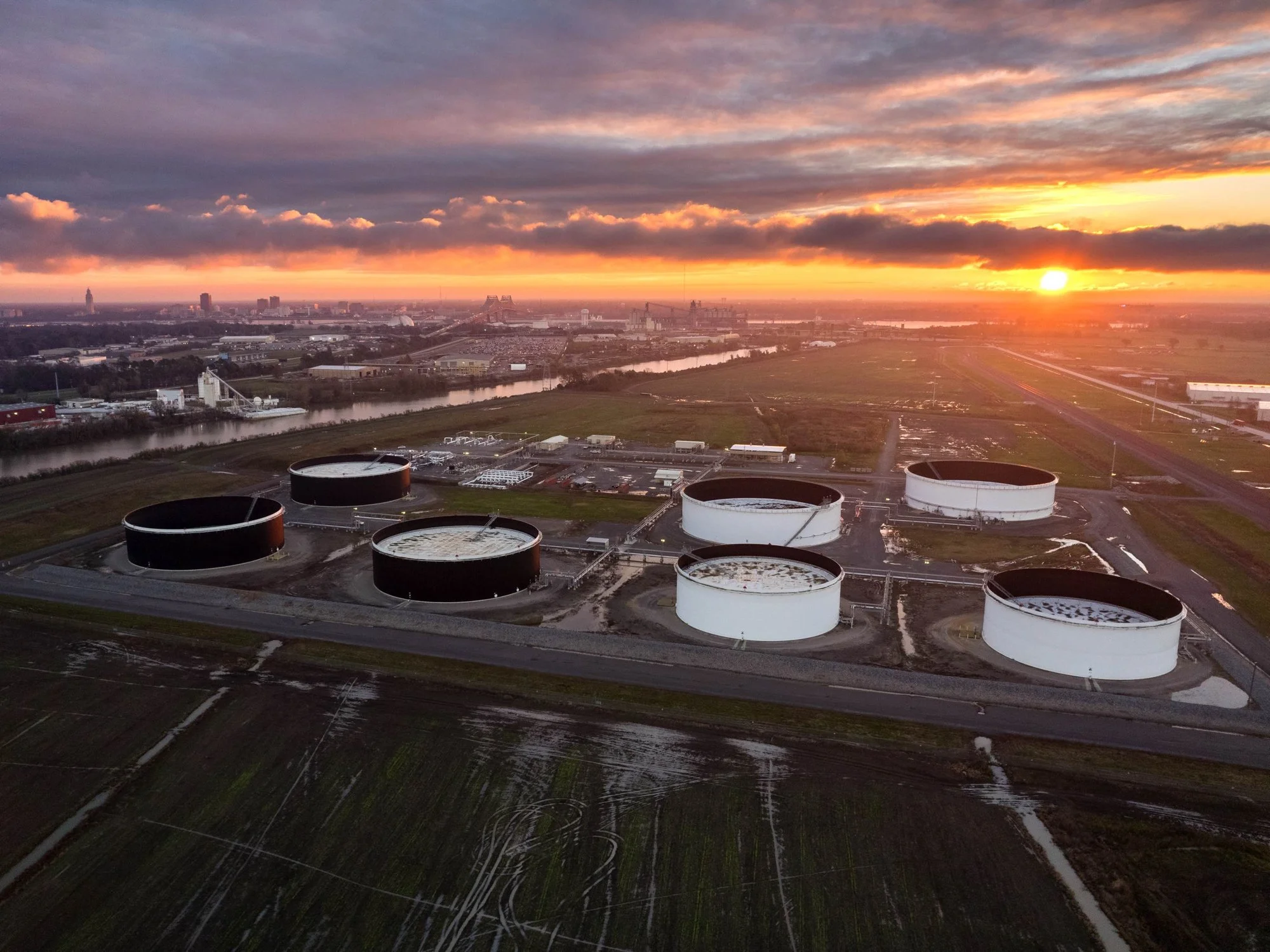 Aerial view of an oil storage facility at sunset with large white and black tanks, a river, and a city in the background.