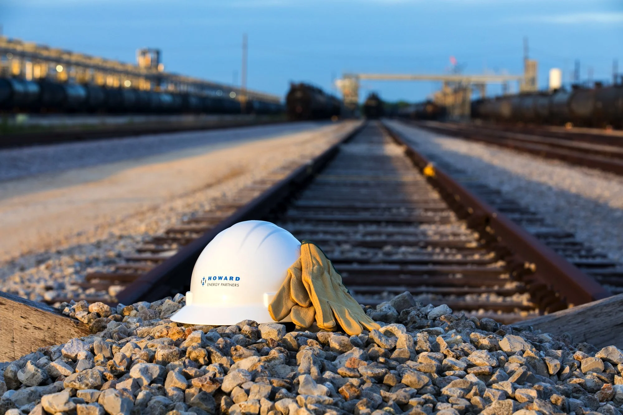 A white safety helmet with the logo 'Howard Energy Partners' and a pair of work gloves resting on gravel next to a railroad track at sunset.