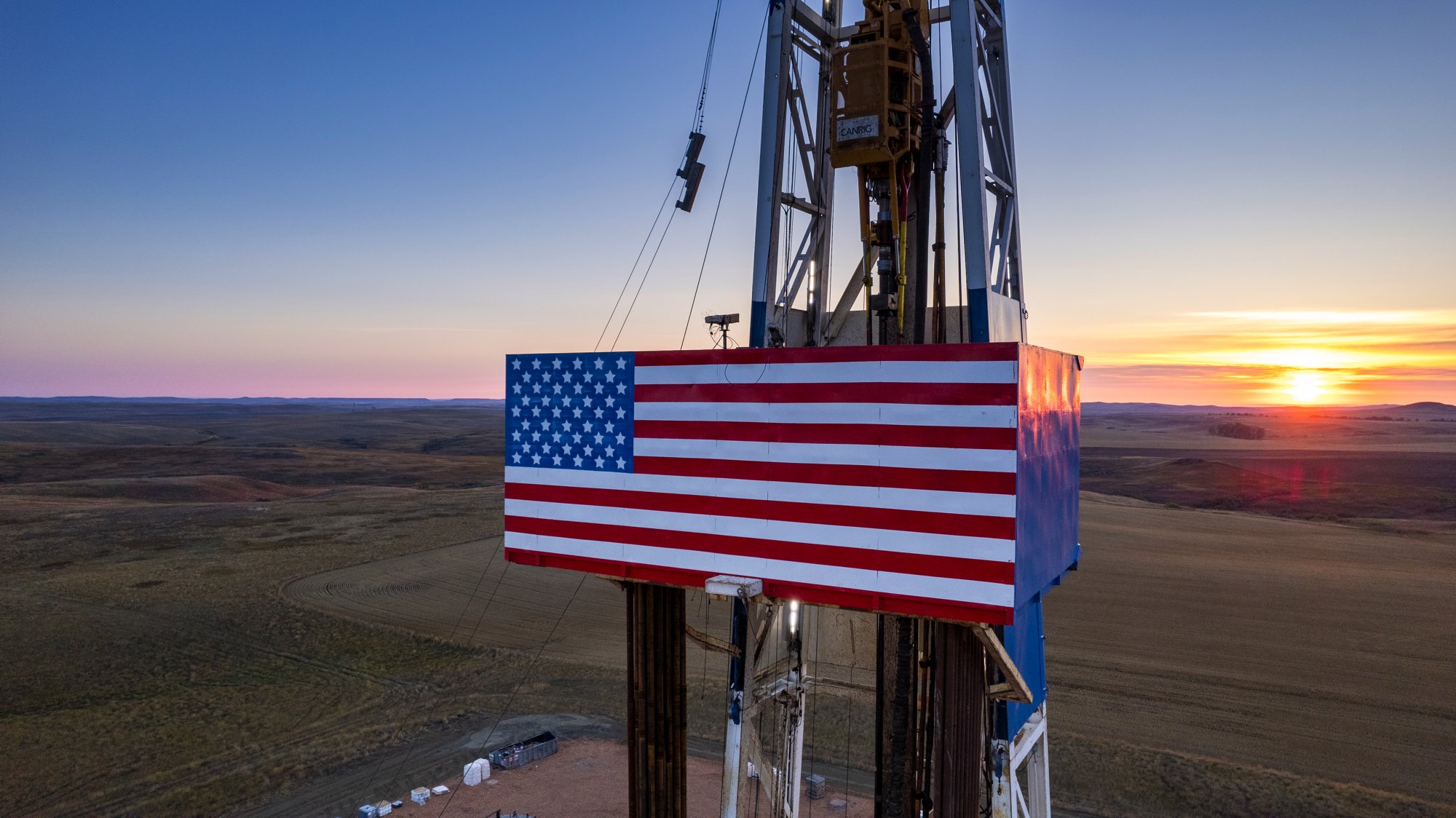An oil rig in a field, decorated with an American flag, during sunset with open plains in the background.