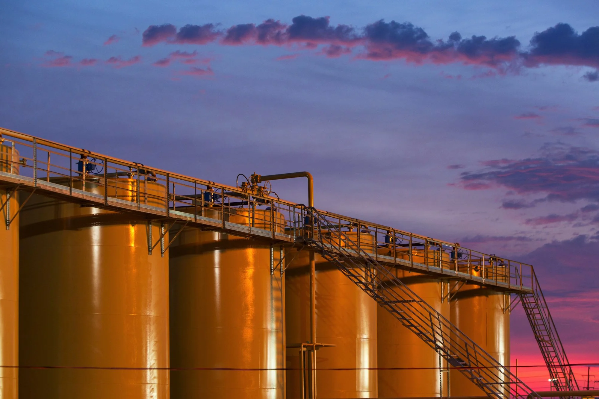 Large industrial storage tanks painted yellow with a staircase, against a colorful sunset sky with clouds.
