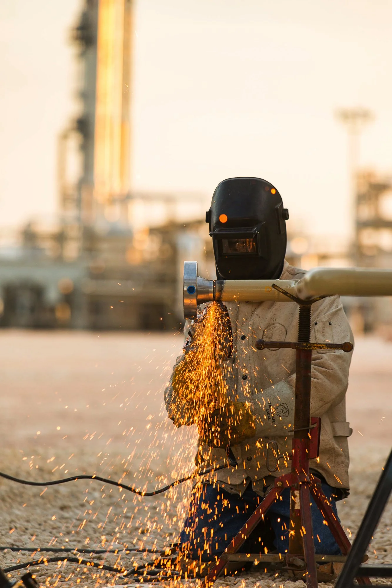 A worker welding metal pipe outdoors during sunset, wearing protective gear, sparks flying.