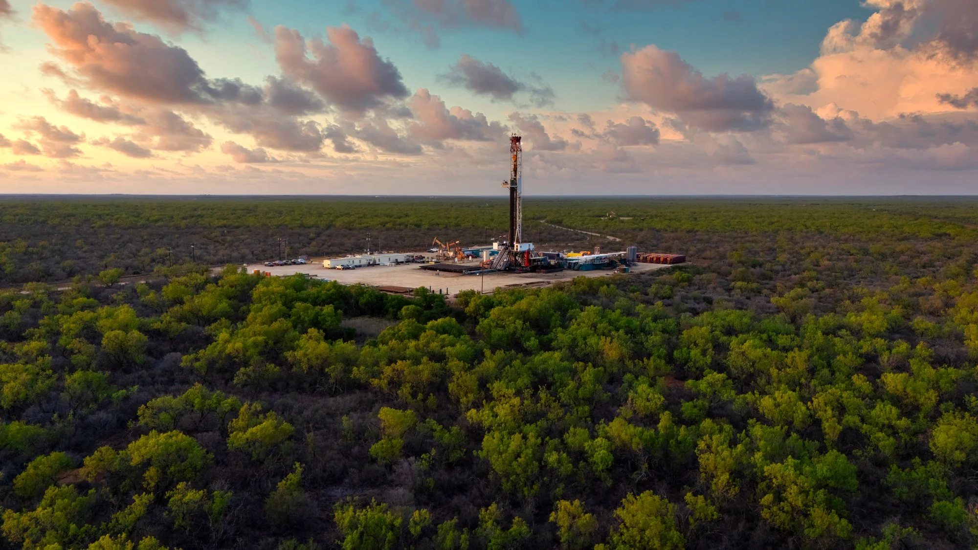 An oil drilling rig in a clearing surrounded by green trees on a flat landscape under a sky with scattered clouds at sunset.