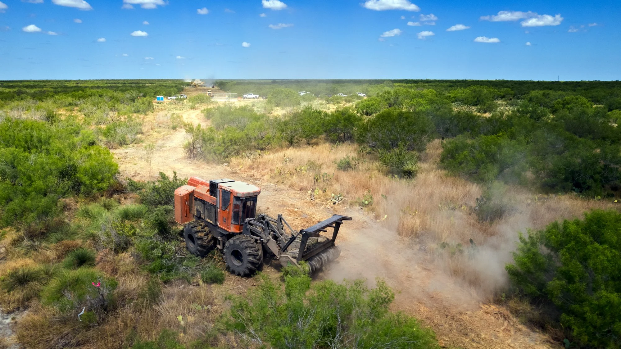 A large orange bulldozer moving dry dirt on a dusty path through sparse desert vegetation, with a clear blue sky and some clouds overhead.