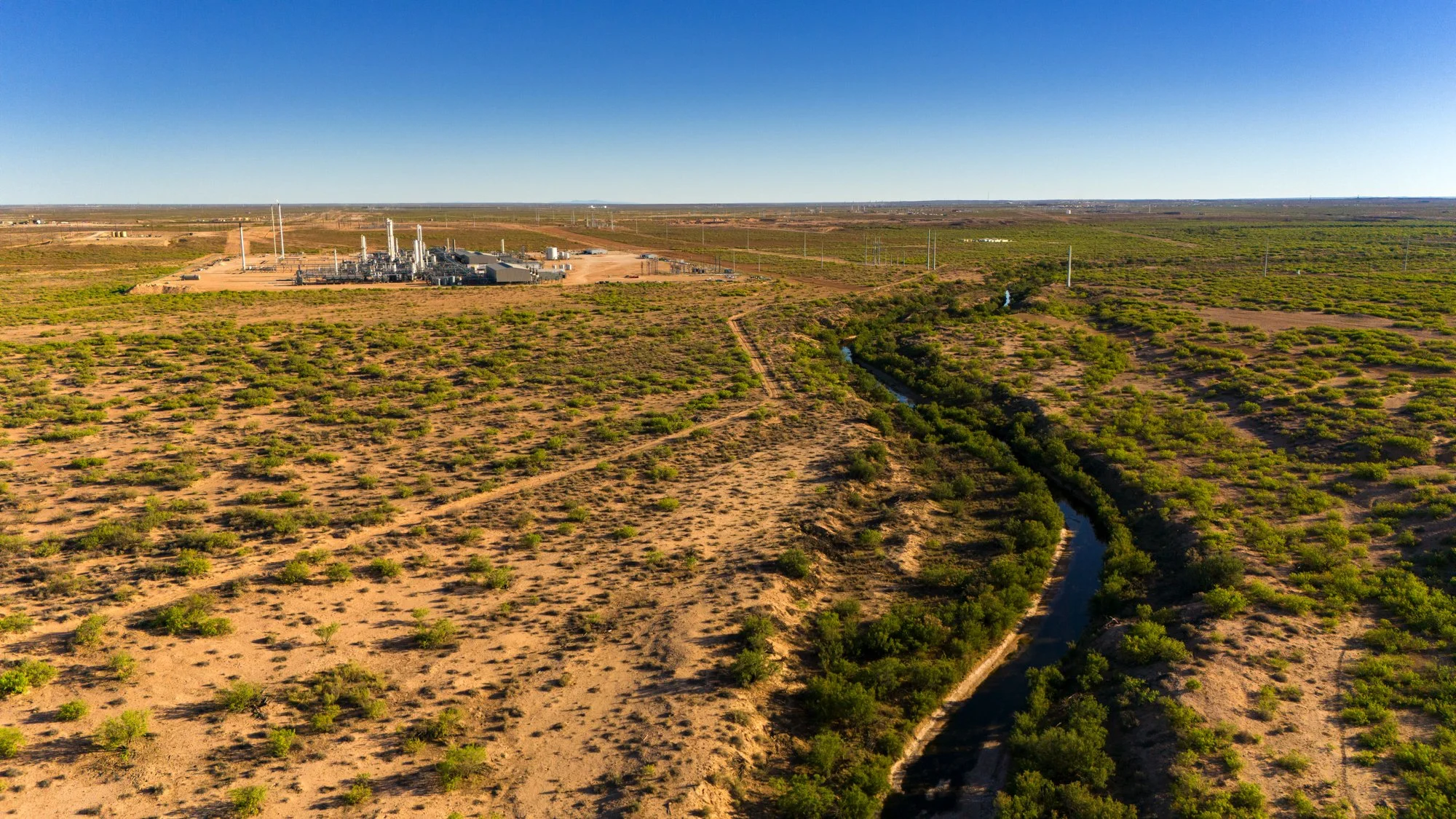 Aerial view of a desert landscape with sparse green vegetation, a narrow winding waterway, and an industrial facility with multiple buildings and smokestacks in the distance under a clear blue sky.