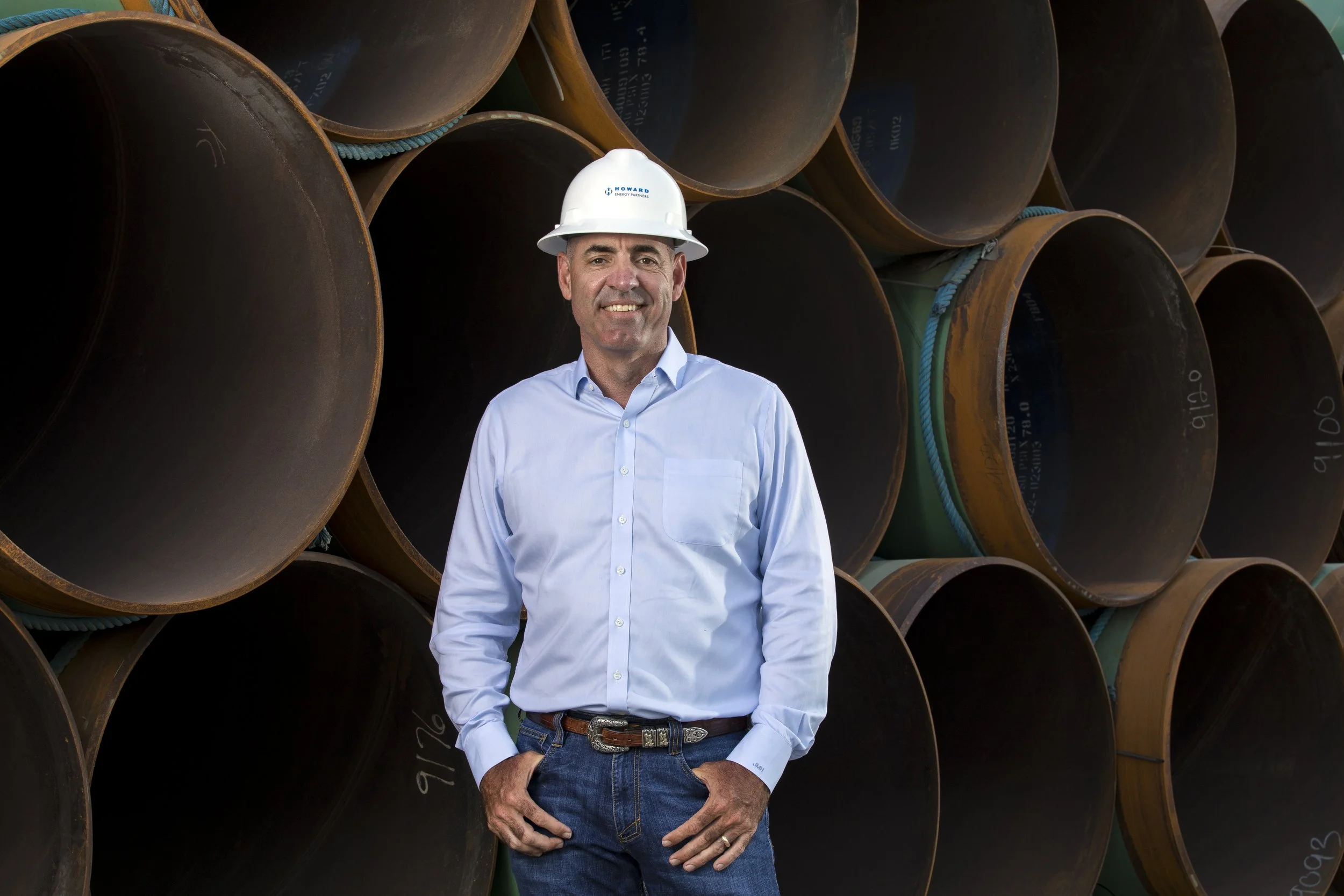 A man in a white hard hat and light blue button-down shirt standing in front of stacked large industrial metal pipes.