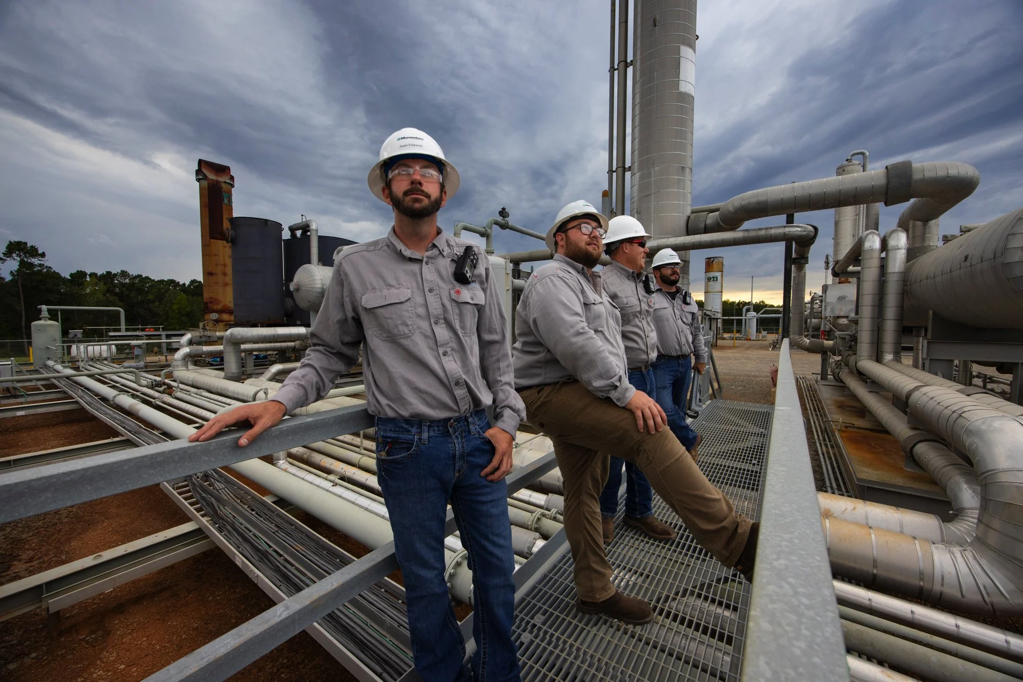 Four workers in gray uniforms and white safety helmets standing on industrial piping platform outdoors under a cloudy sky.