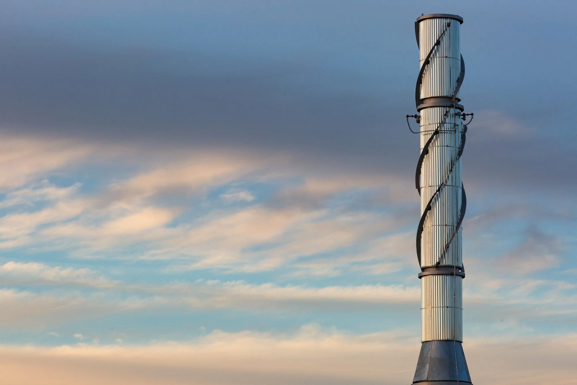 A tall, modern industrial chimney with a spiral metal ladder wrapped around it, set against a sky with scattered clouds.
