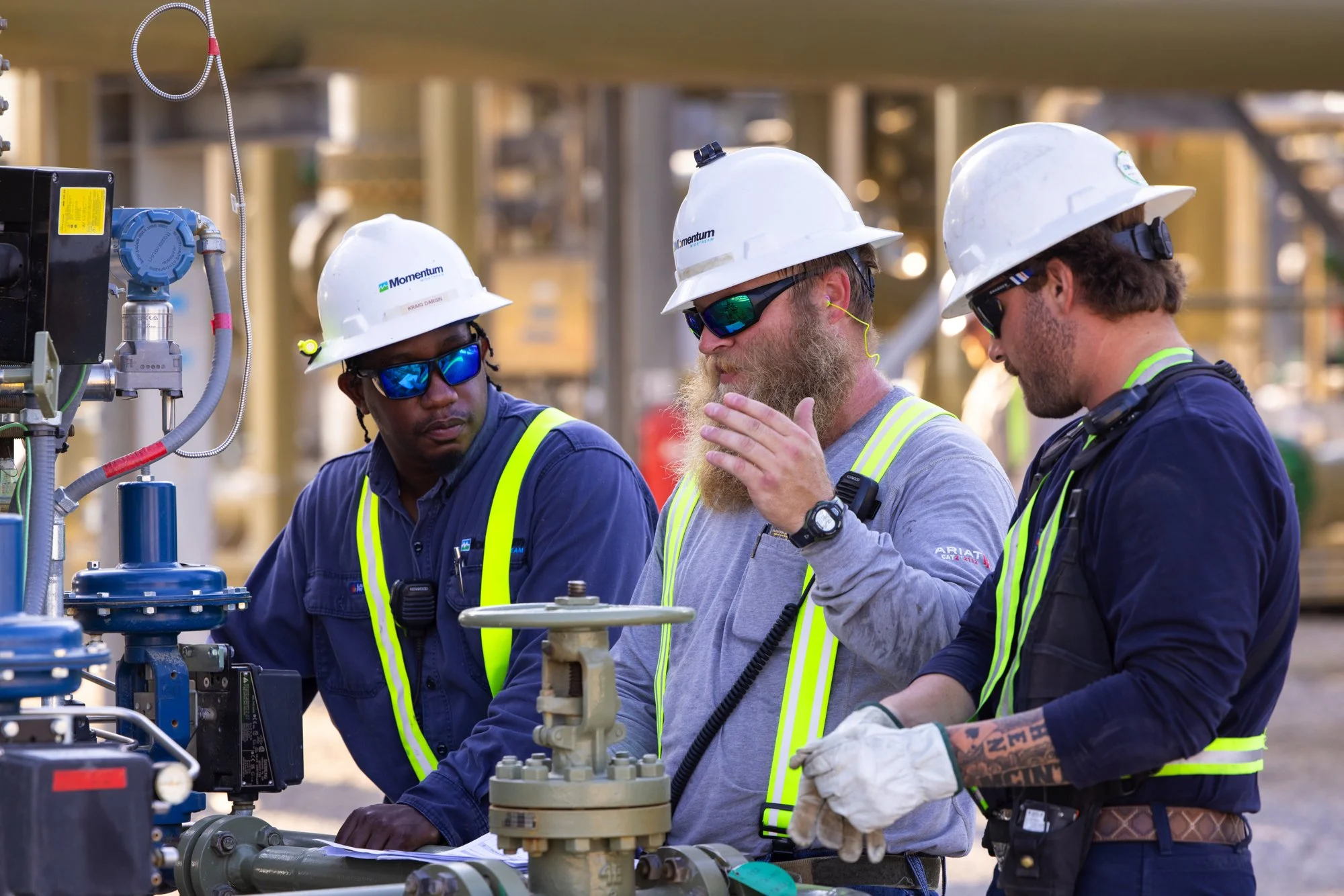 Three construction workers wearing white hard hats, safety glasses, and reflective vests, discussing while inspecting equipment outdoors at a construction site.