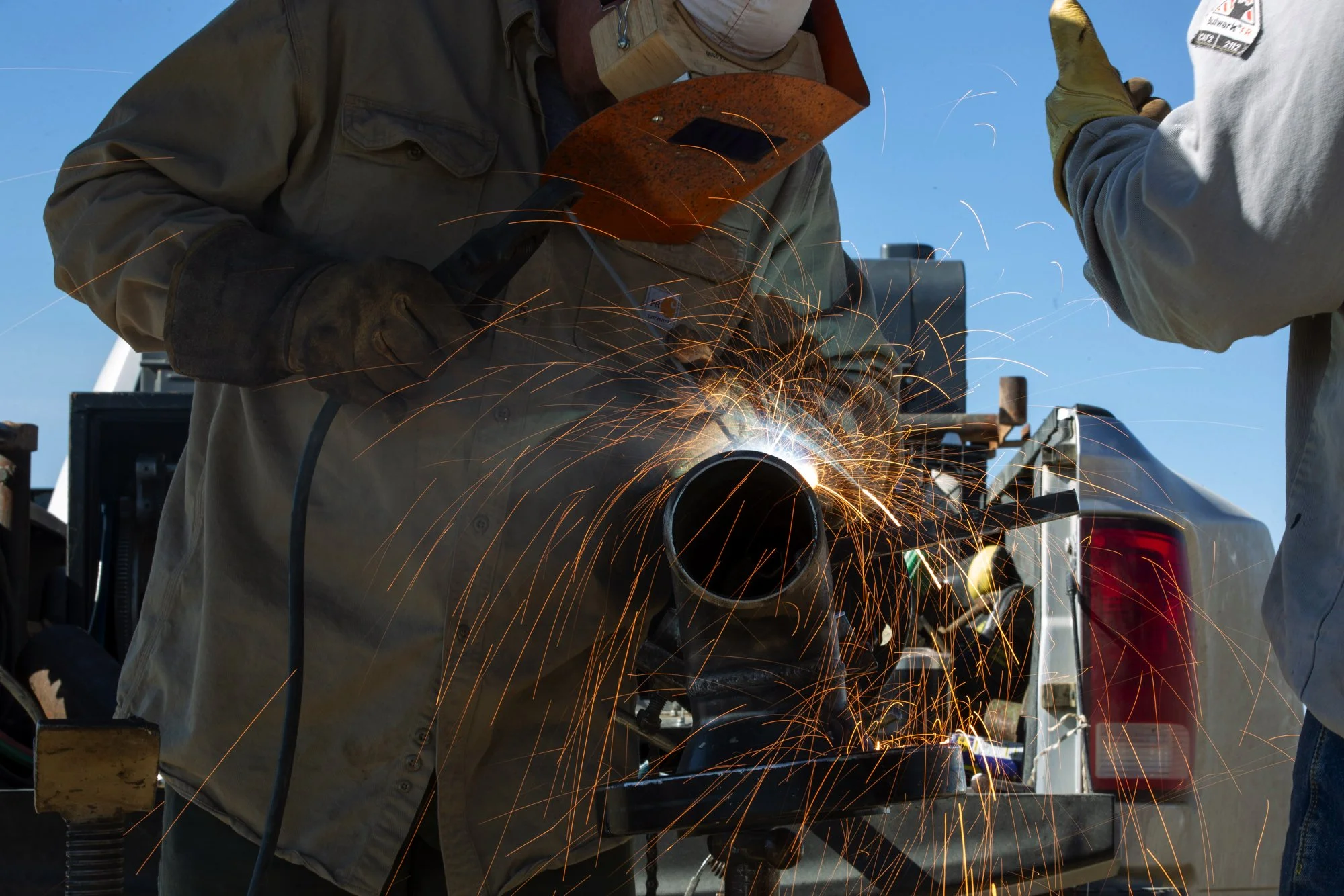 Two workers welding a metal pipe with sparks flying, outdoors on a sunny day.