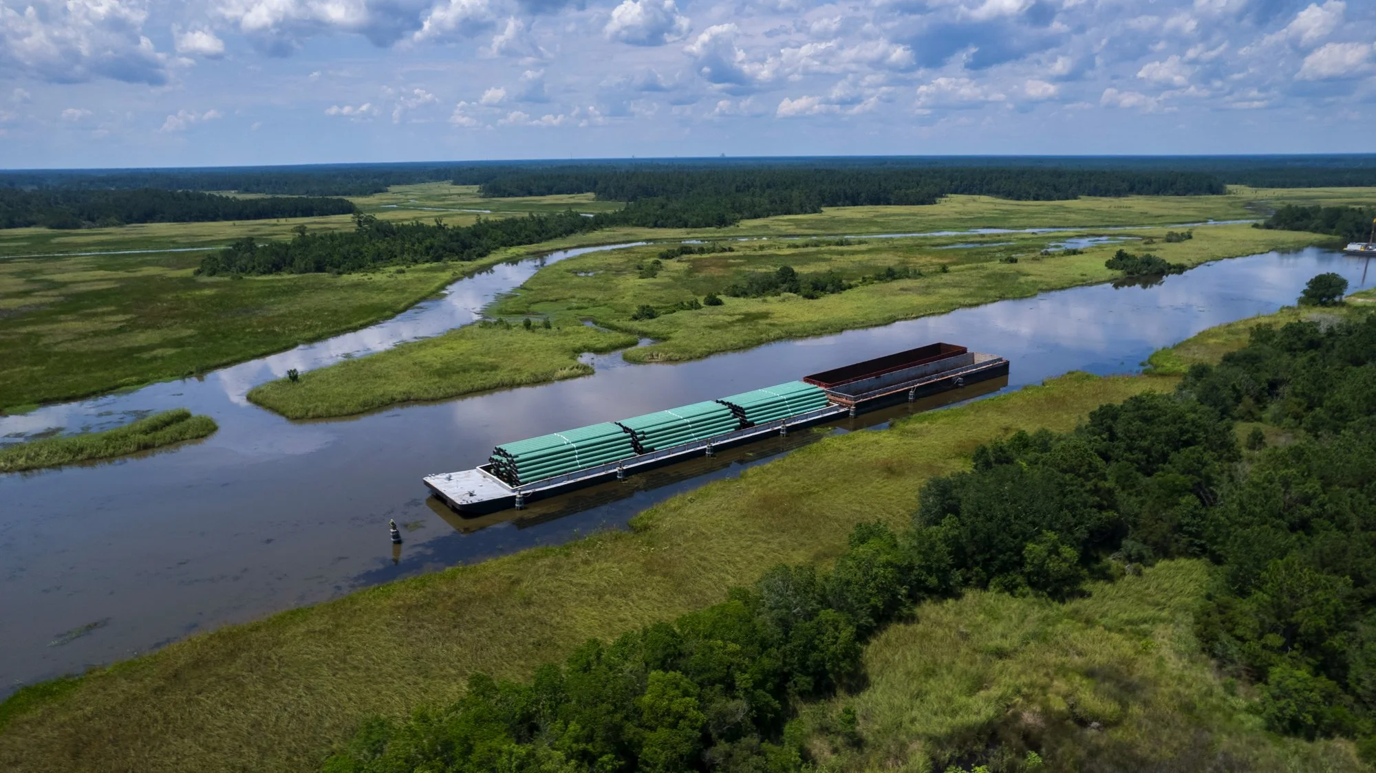 A barge carrying large pipes on water surrounded by green wetlands and trees under a partly cloudy sky.