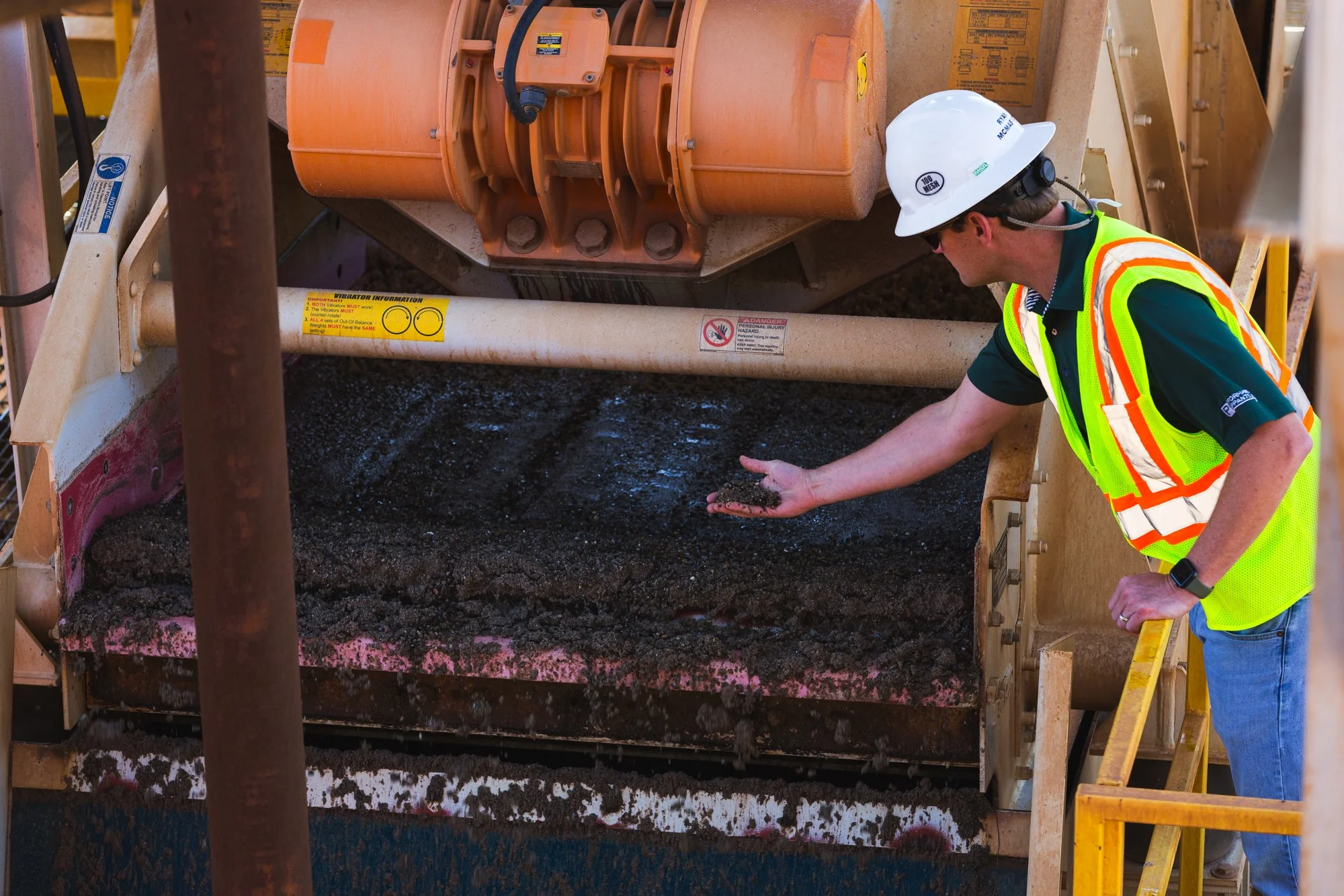 A construction worker wearing a white hard hat, sunglasses, and a yellow safety vest is inspecting and holding a handful of dark asphalt or gravel at a construction site, with heavy machinery and construction equipment in the background.