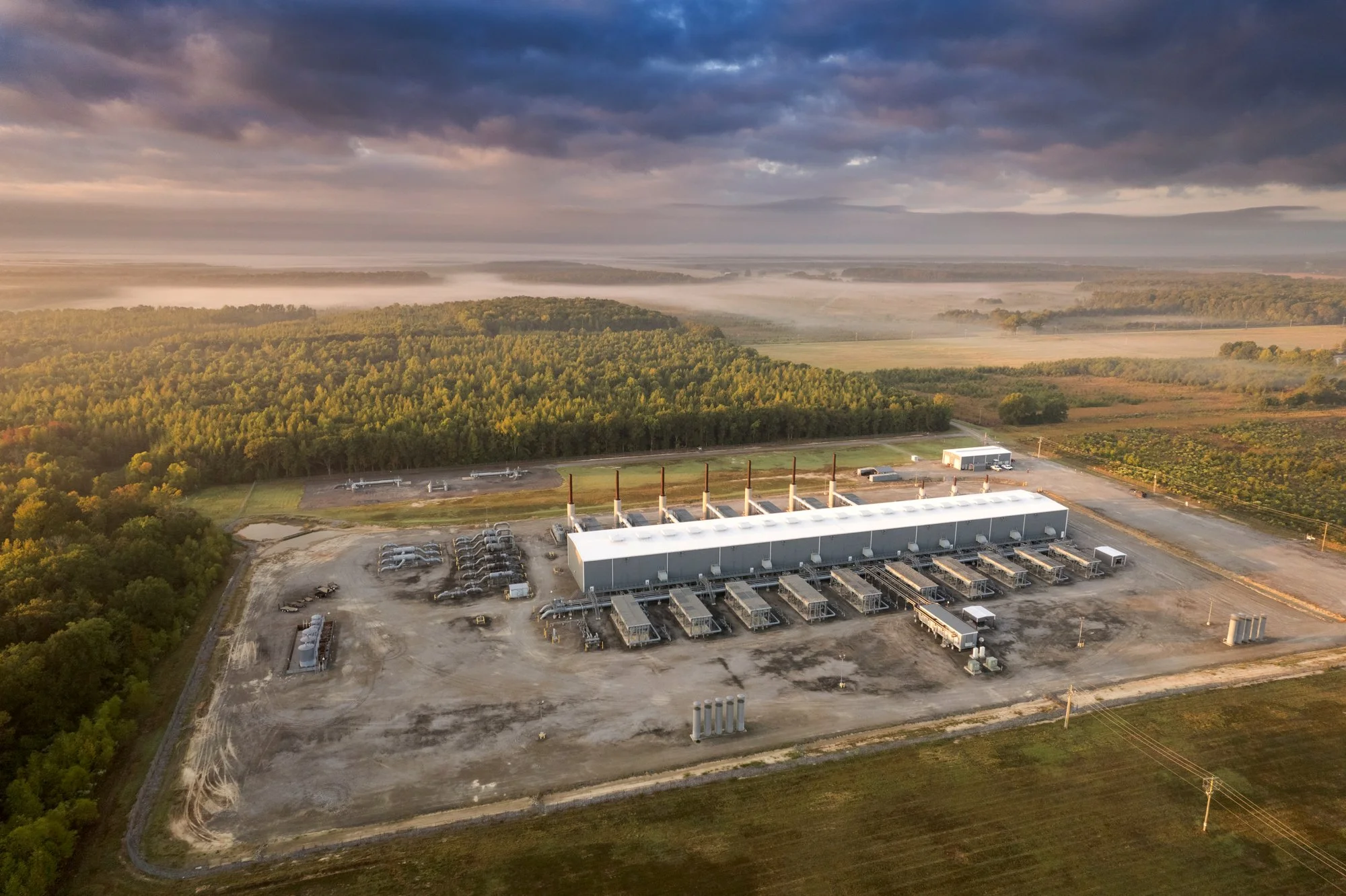 Aerial view of a large industrial facility with multiple smokestacks, surrounded by farmland and a forest, under a cloudy sky.