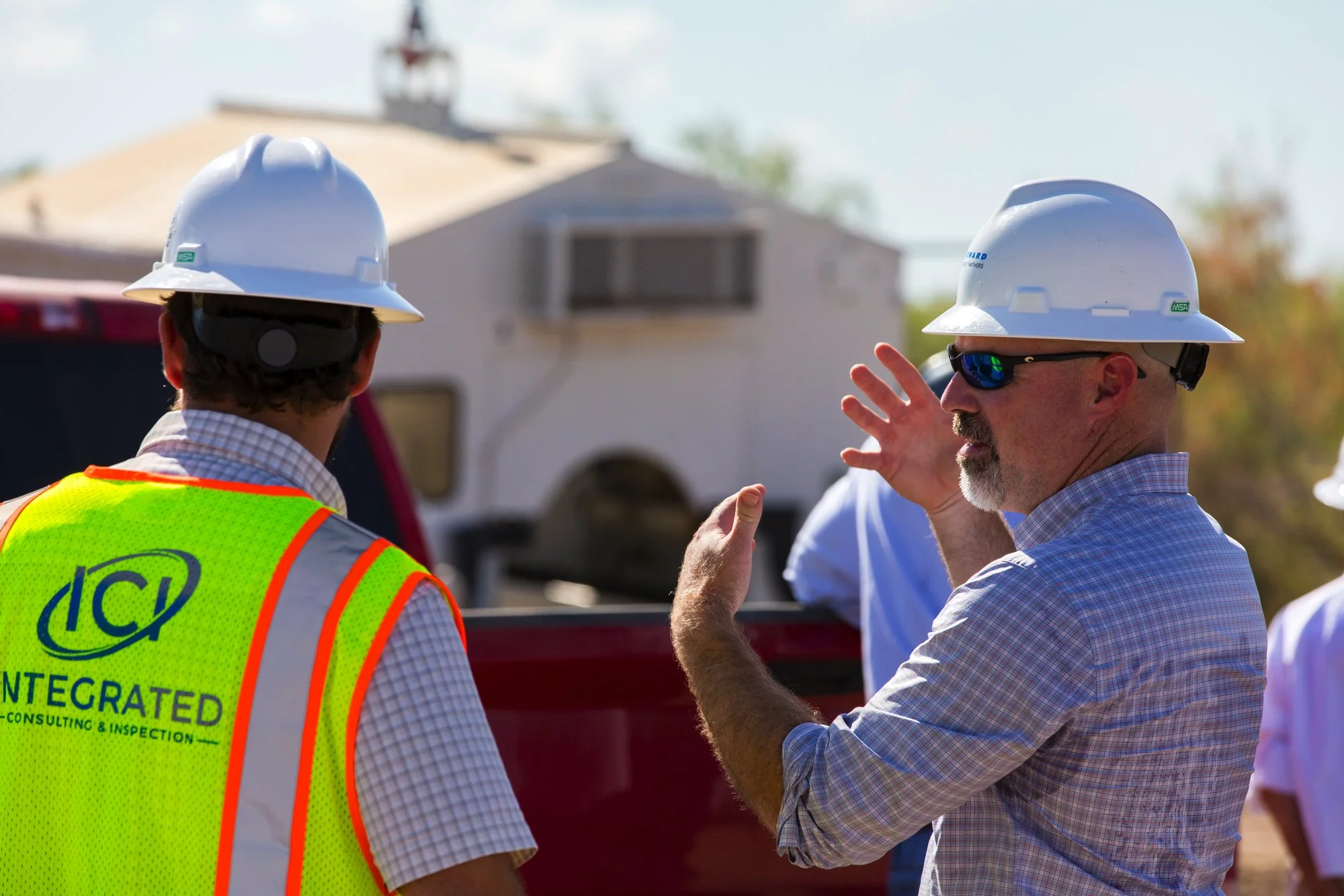Two construction workers wearing white safety helmets and reflective vests, engaged in conversation outdoors, with a building and construction vehicle in the background.