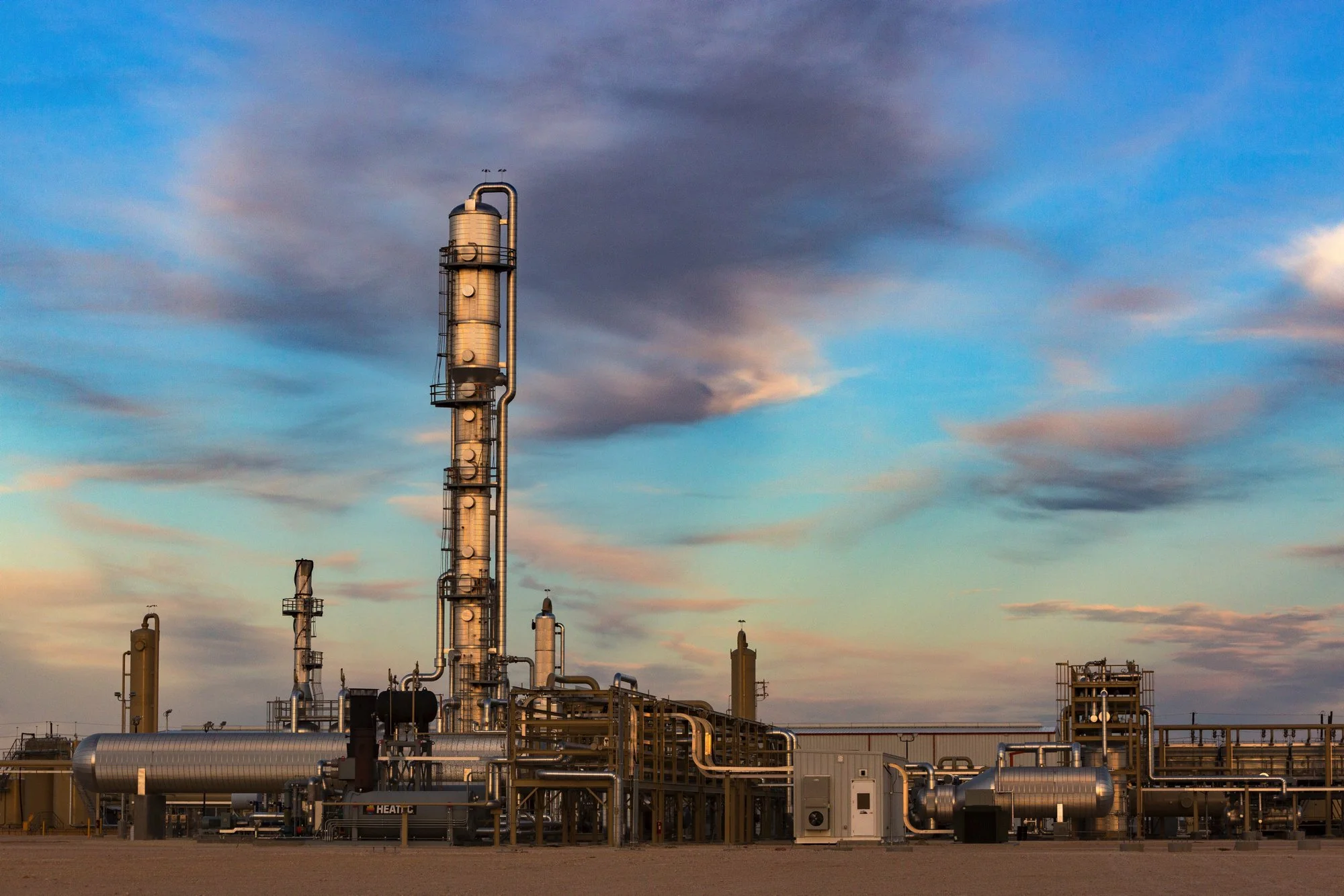 A large industrial refinery with tall vertical processing towers and extensive pipes and machinery, set against a partly cloudy sky during sunset.