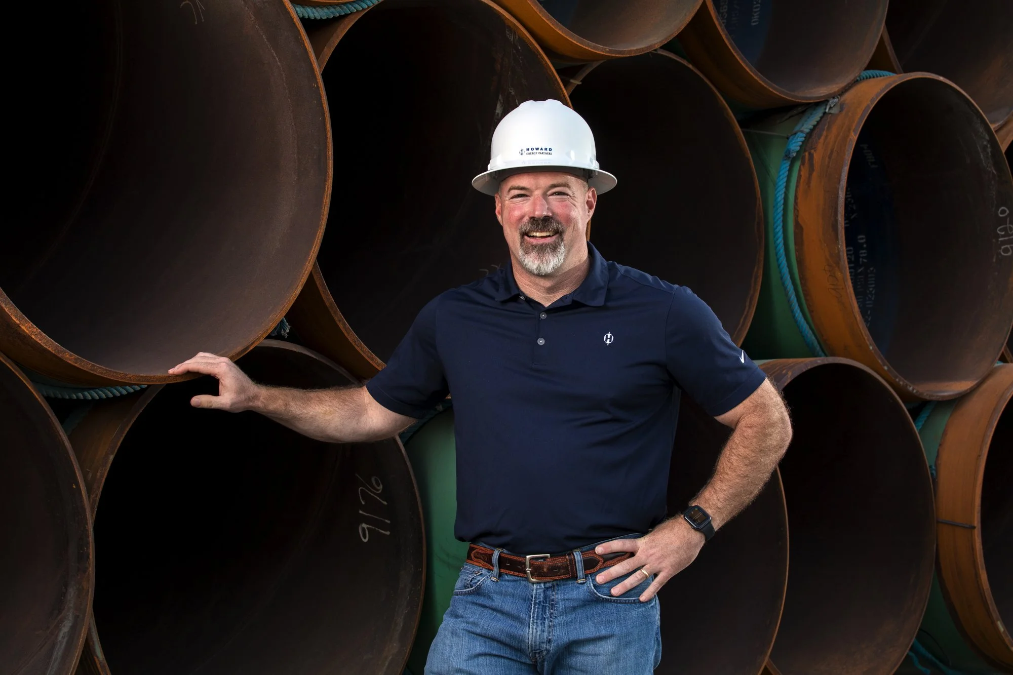 A man in a white safety helmet and navy polo shirt smiling at the camera, standing among large stacked pipes at an industrial site.