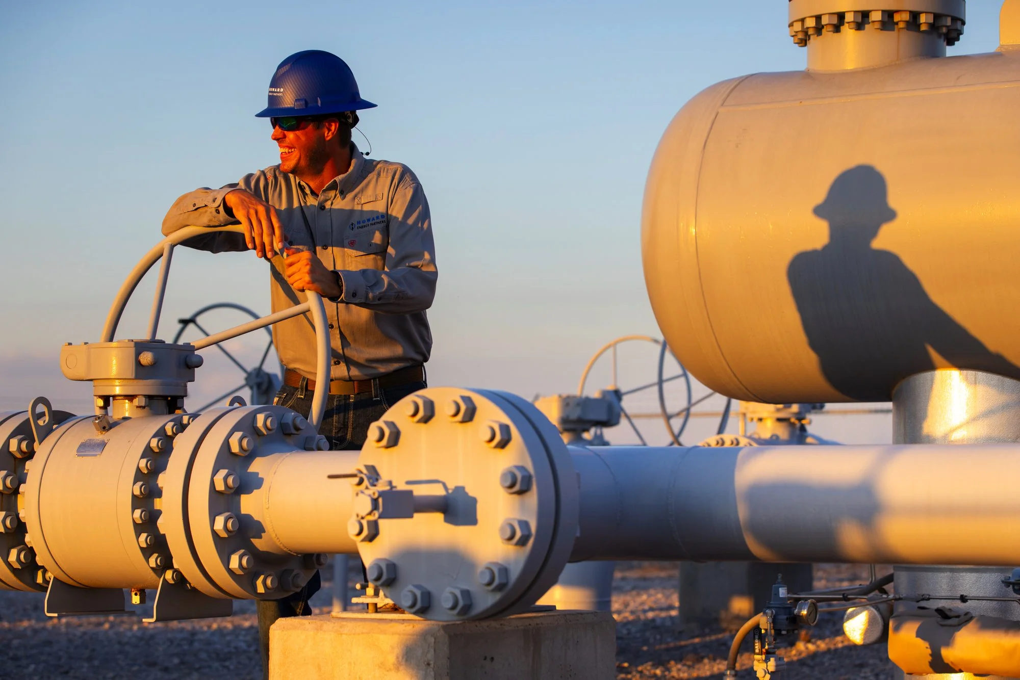 A worker in safety gear smiling and looking away while standing next to industrial pipes and equipment at an oil or gas facility during sunset.