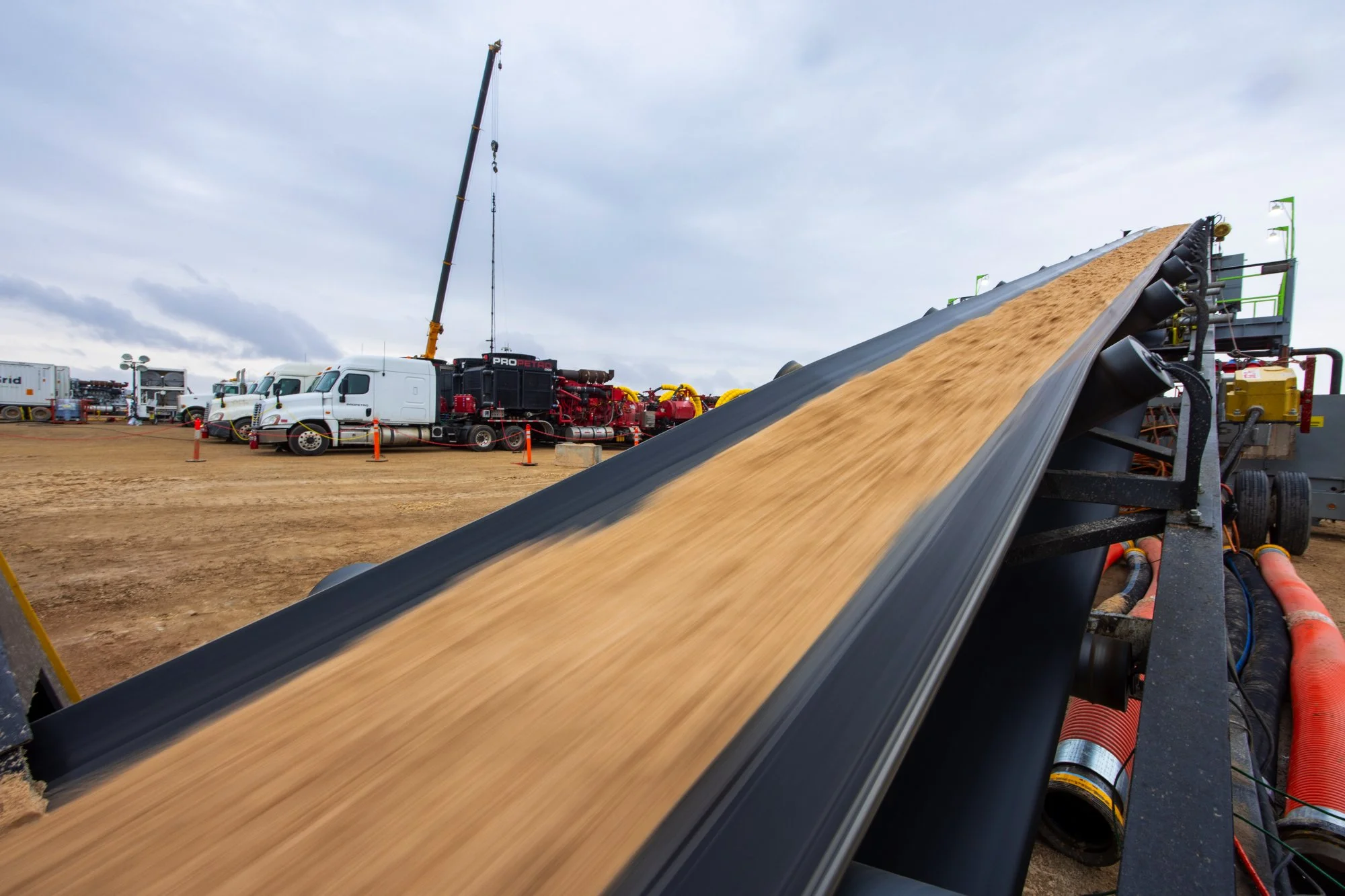 Conveyor belt transporting sand on a construction site with trucks and construction equipment in the background under cloudy sky.