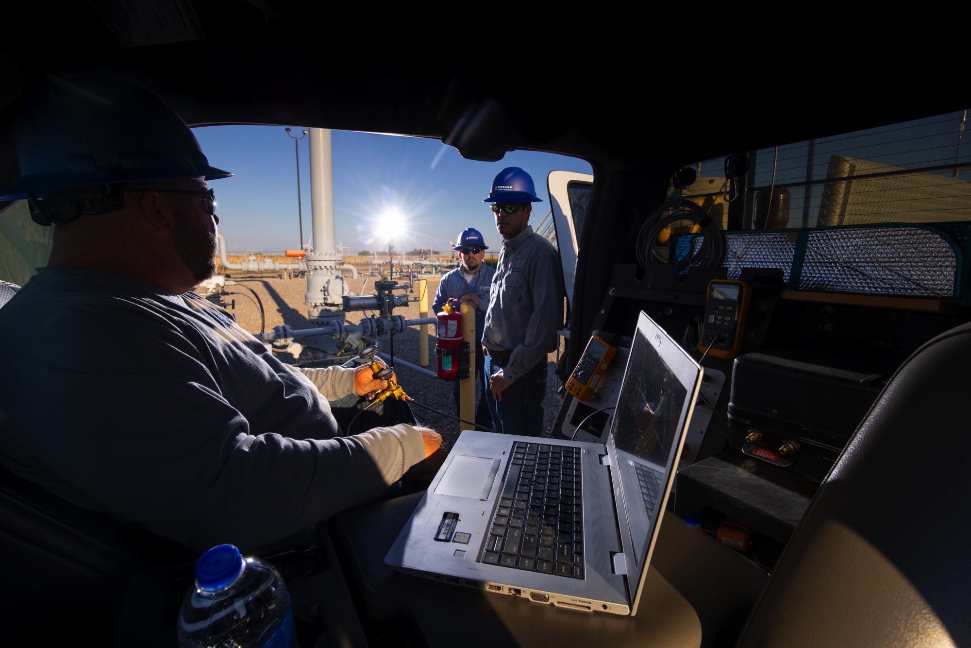 Workers wearing safety helmets and sunglasses standing near oil or gas pipelines in a desert environment during sunset or sunrise, with one worker inside a control vehicle operating equipment and others outside observing.