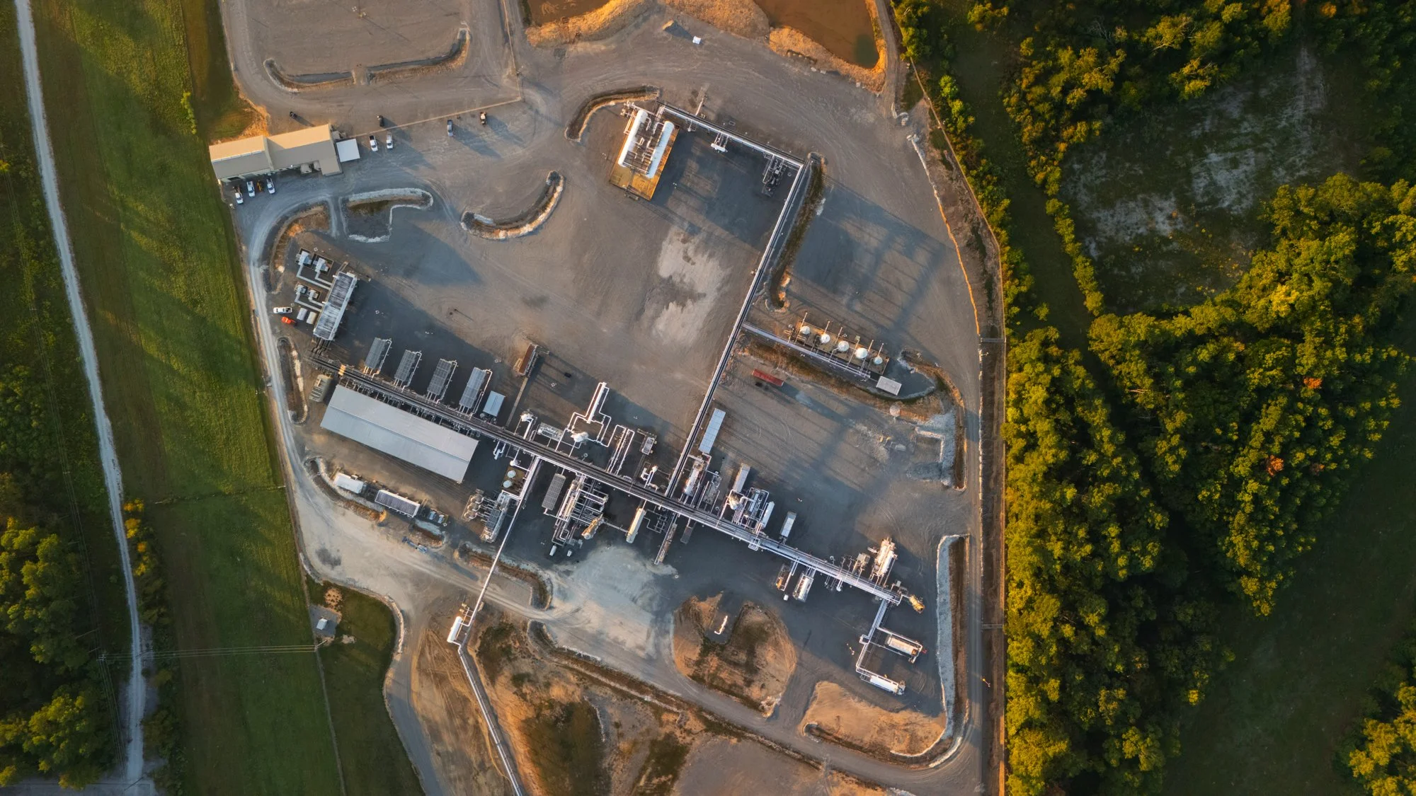 Aerial view of an industrial facility surrounded by green trees and open land, with various buildings, pipelines, and equipment visible.