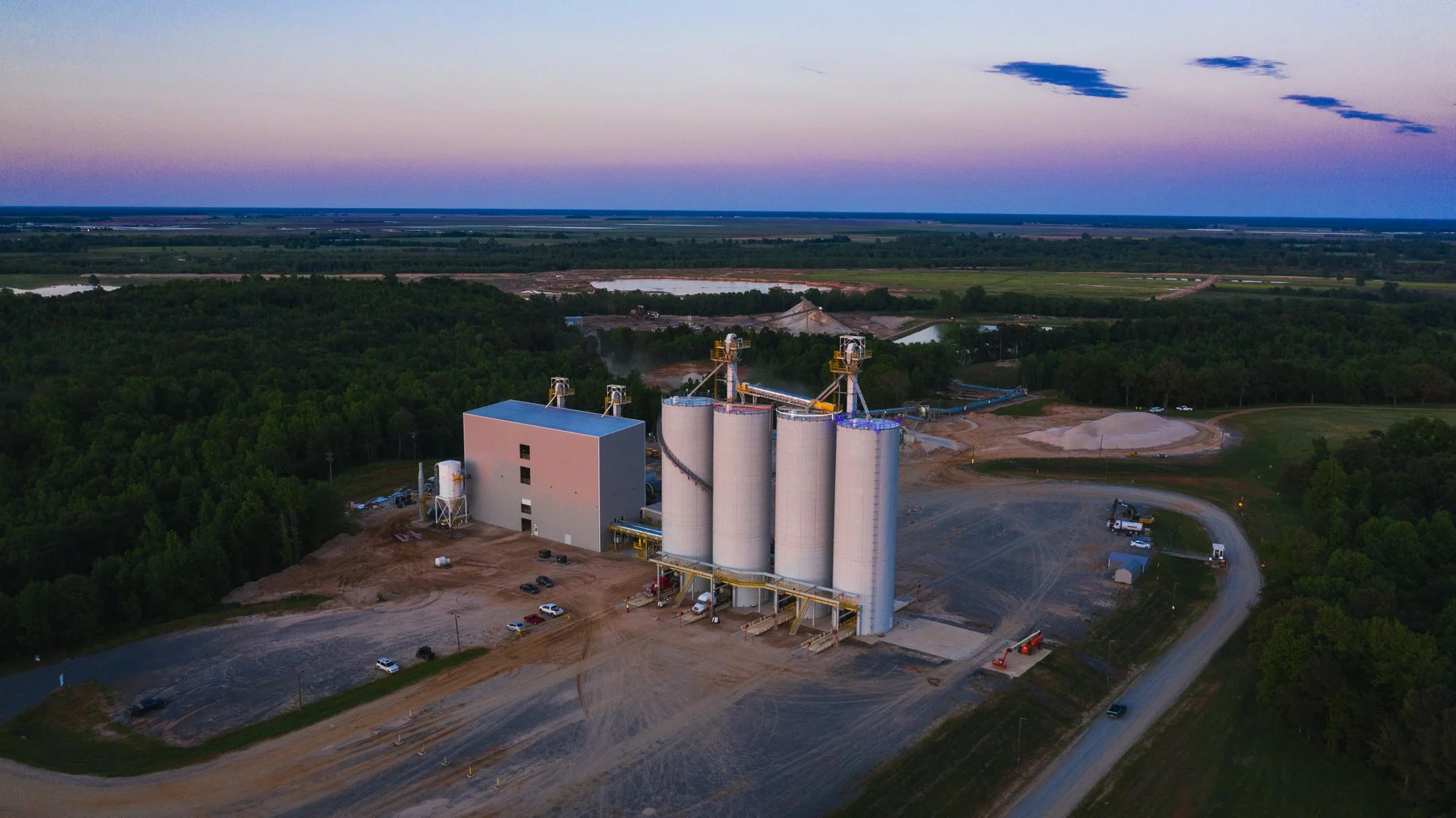 Aerial view of an industrial facility with four large white silos, a building, and a dirt road, surrounded by greenery and open land at dusk.
